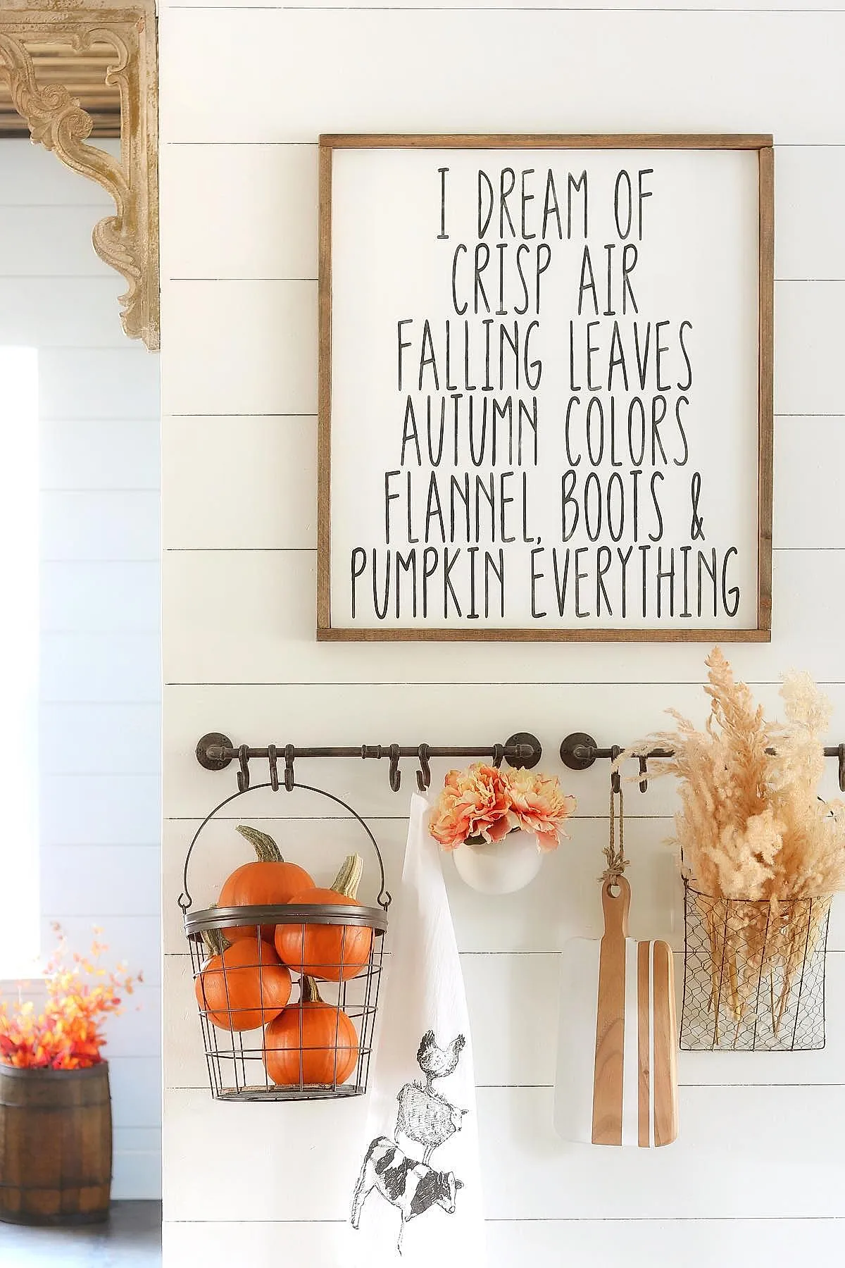 Wire basket filled with small orange pumpkins hanging on black metal hooks against white shiplap wall, alongside a white towel with farm animal prints, pink dried flowers in a round white vase, wooden cutting boards, and dried grasses in a wire container.