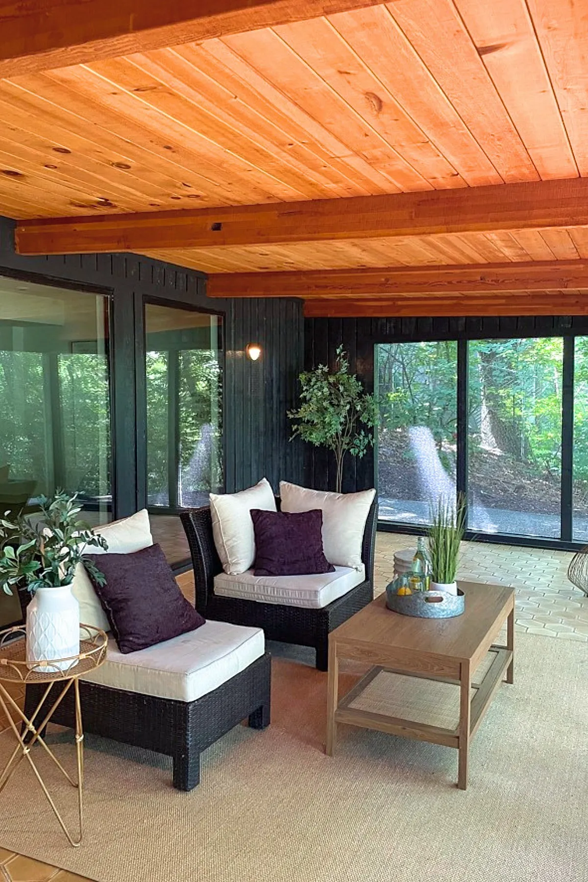 Screened in porch with warm natural wood ceiling, dark wicker chairs with cream cushions and plum pillows, wooden coffee table with plants, large sliding glass windows showing green trees outside