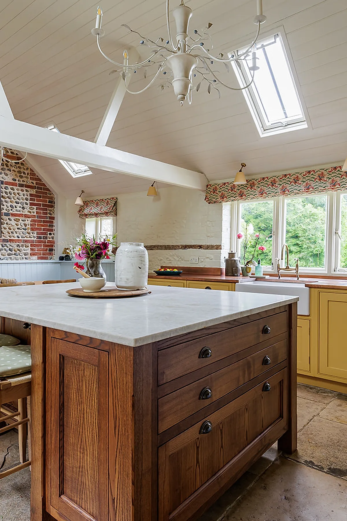Rustic kitchen featuring a large wooden island with marble countertop, yellow lower cabinets with copper countertops, floral window valances, and exposed brick and stone wall under white painted vaulted ceiling with skylights.