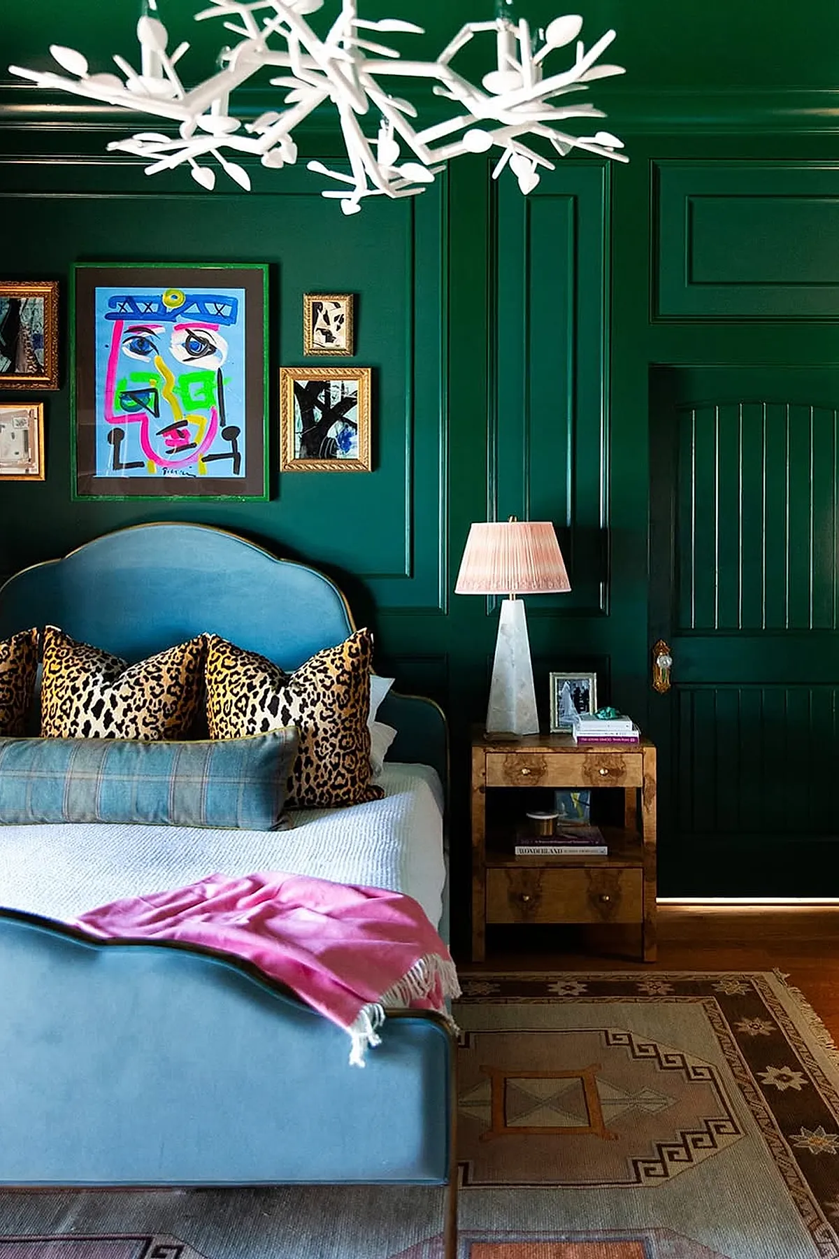 Dark feminine bedroom with deep green paneled walls, light blue upholstered bed with gold trim, leopard print pillows, plaid bolster pillow, pink throw blanket, wooden nightstand with white lamp, and colorful abstract art above the headboard.