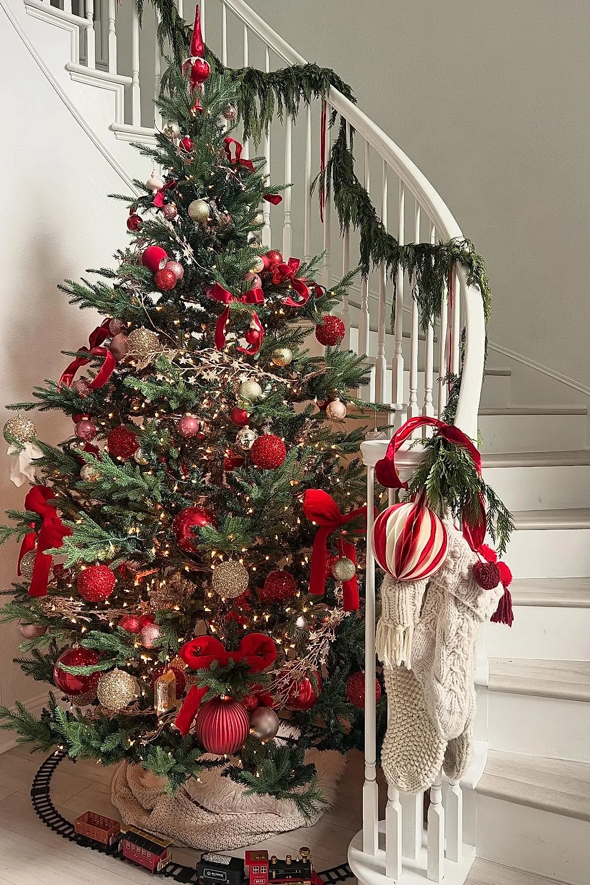 Christmas tree decorated with red and gold ornaments, velvet ribbons, surrounded by knitted stockings hanging on white staircase banister, green garland draped on railing, toy train circling tree base