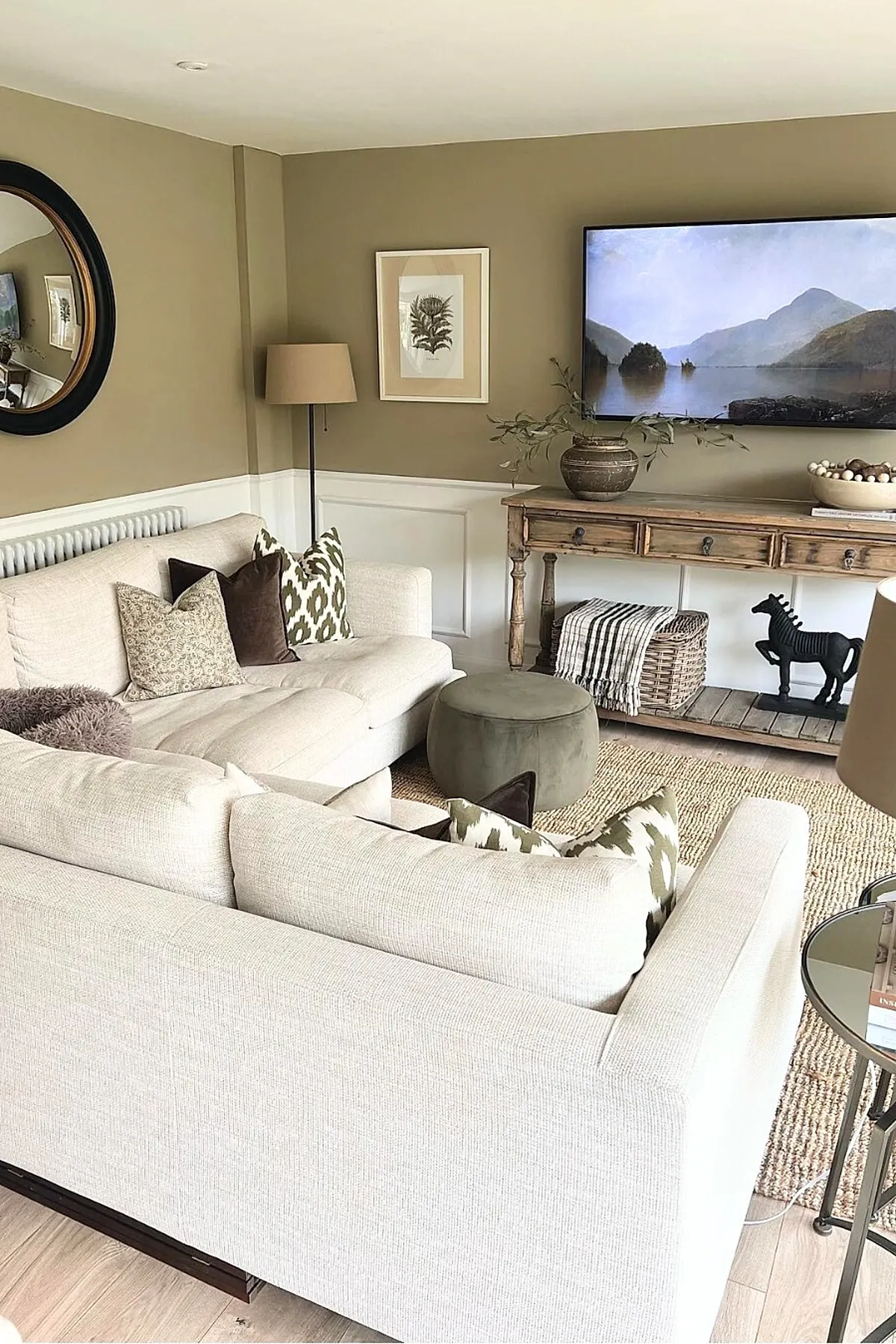 Earthy living room with beige sectional sofas adorned with patterned and solid throw pillows in shades of brown and green, a round olive green ottoman, a wooden console table with drawers displaying a ceramic vase with branches and decorative bowls. The walls are painted warm taupe above white wainscoting, featuring a round black-framed mirror, botanical wall art, and a large flat-screen TV showing a nature scene. The floor is covered with a woven jute rug.