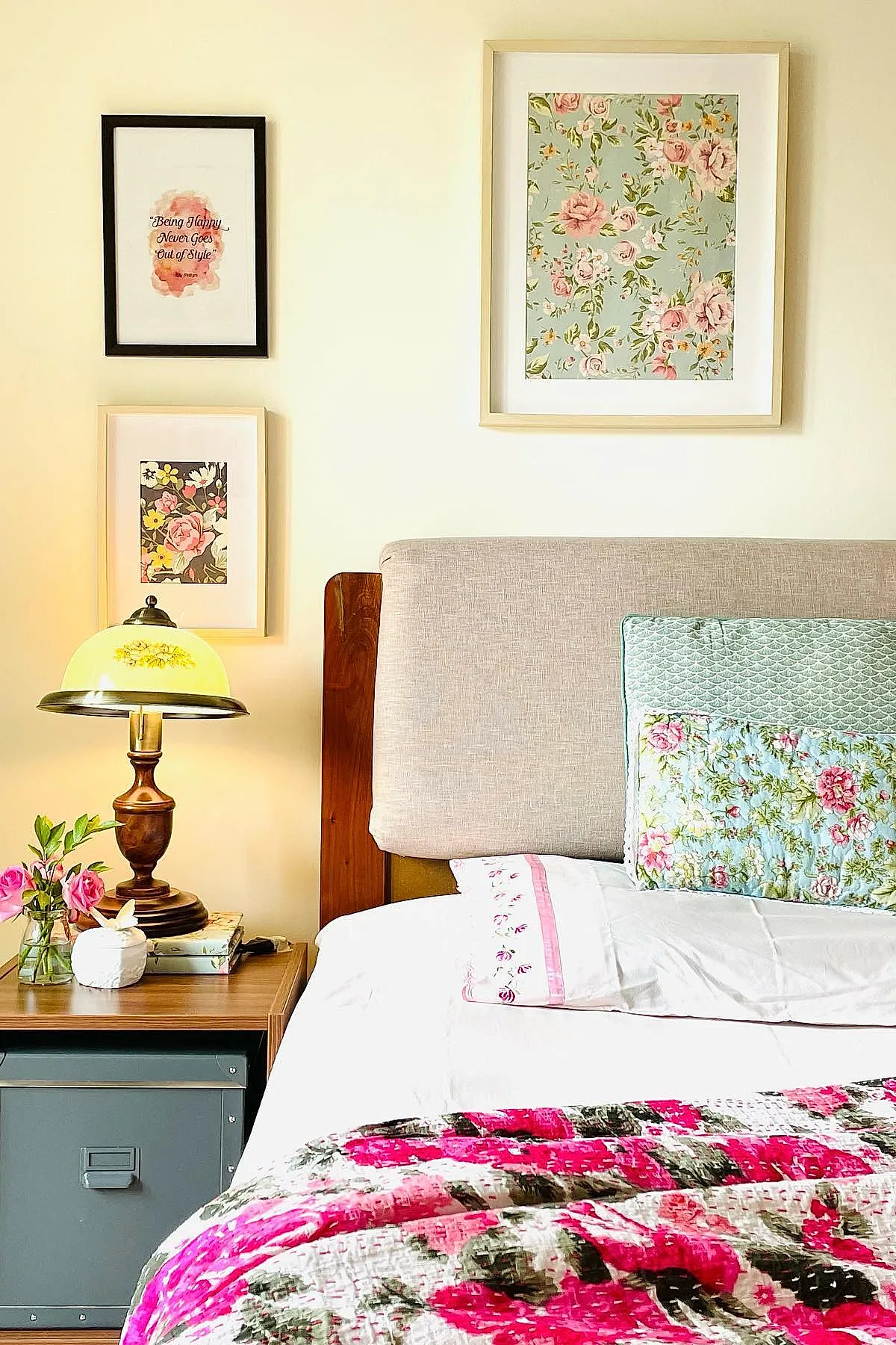 bedside corner featuring beige upholstered headboard, bed dressed with bright pink floral quilt and pastel floral pillows, wooden nightstand holding antique brass lamp with yellow glass shade, small vase of pink roses, three framed artworks on cream wall
