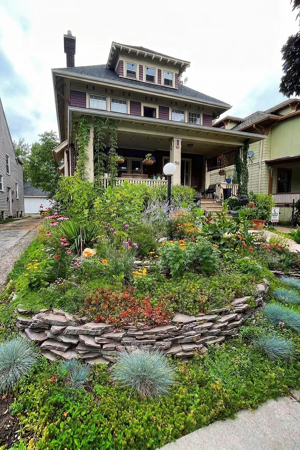 Small front yard landscaping featuring layered flower beds with colorful plants including pink, purple, yellow, and orange flowers, supported by a rustic stacked stone retaining wall, lush green ground cover, and climbing vines on the porch of a two-story house with beige columns and purple siding.