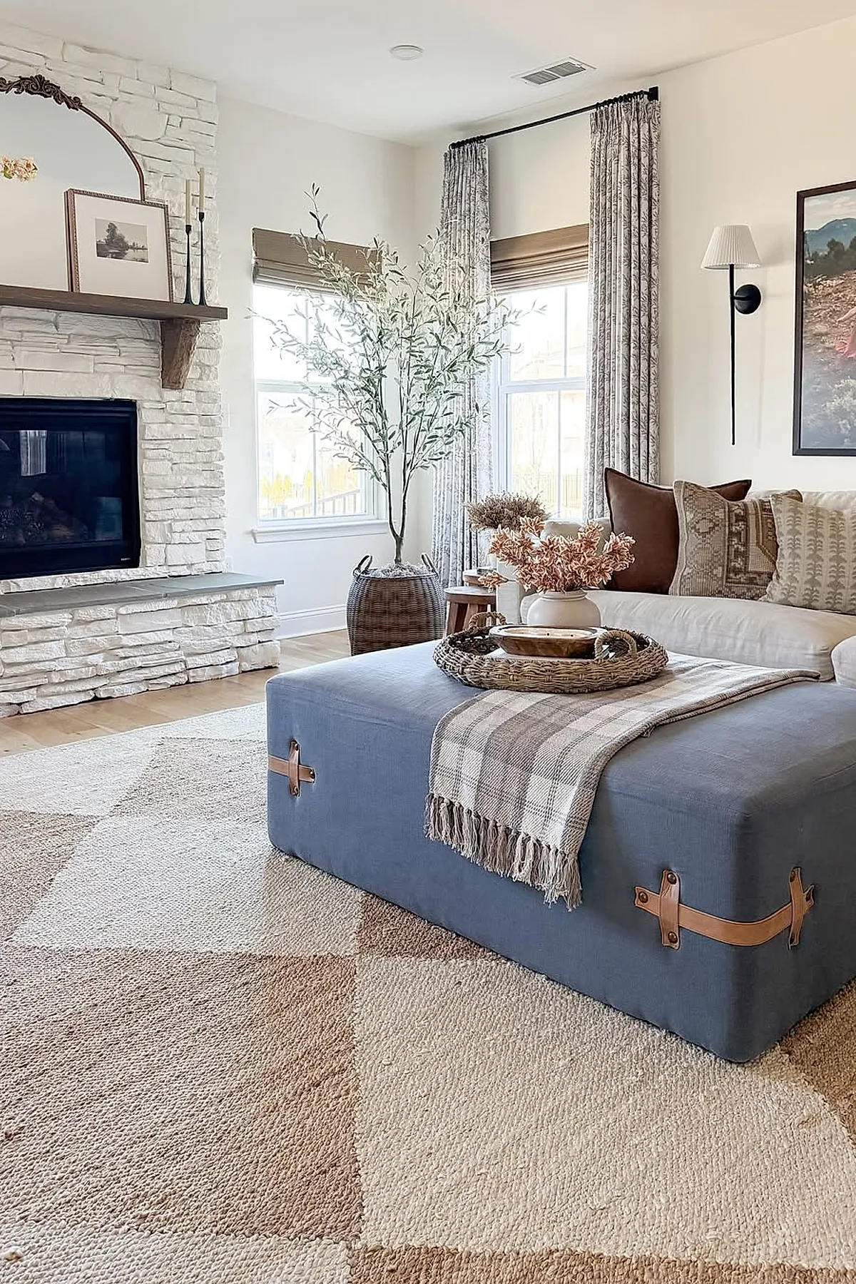 Earthy living room featuring a large blue fabric ottoman with leather handles topped with a plaid throw and wicker tray holding dried flowers. The space includes a white stone fireplace with wooden mantel, tall windows dressed in patterned curtains and woven shades, neutral-colored sofa with brown and patterned pillows, light wood floors, and a geometric beige and brown rug.