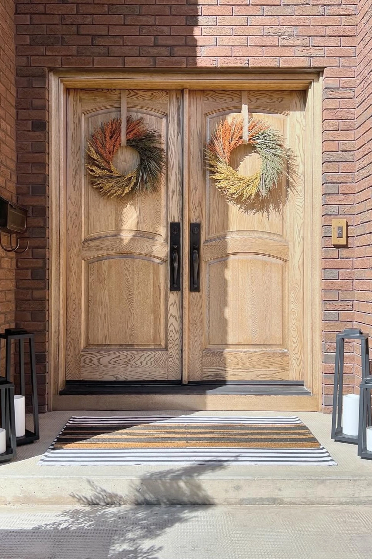Oak double front doors with matching fall grass wreaths, black lantern lanterns with candles, and a layered striped doormat against a brick entry