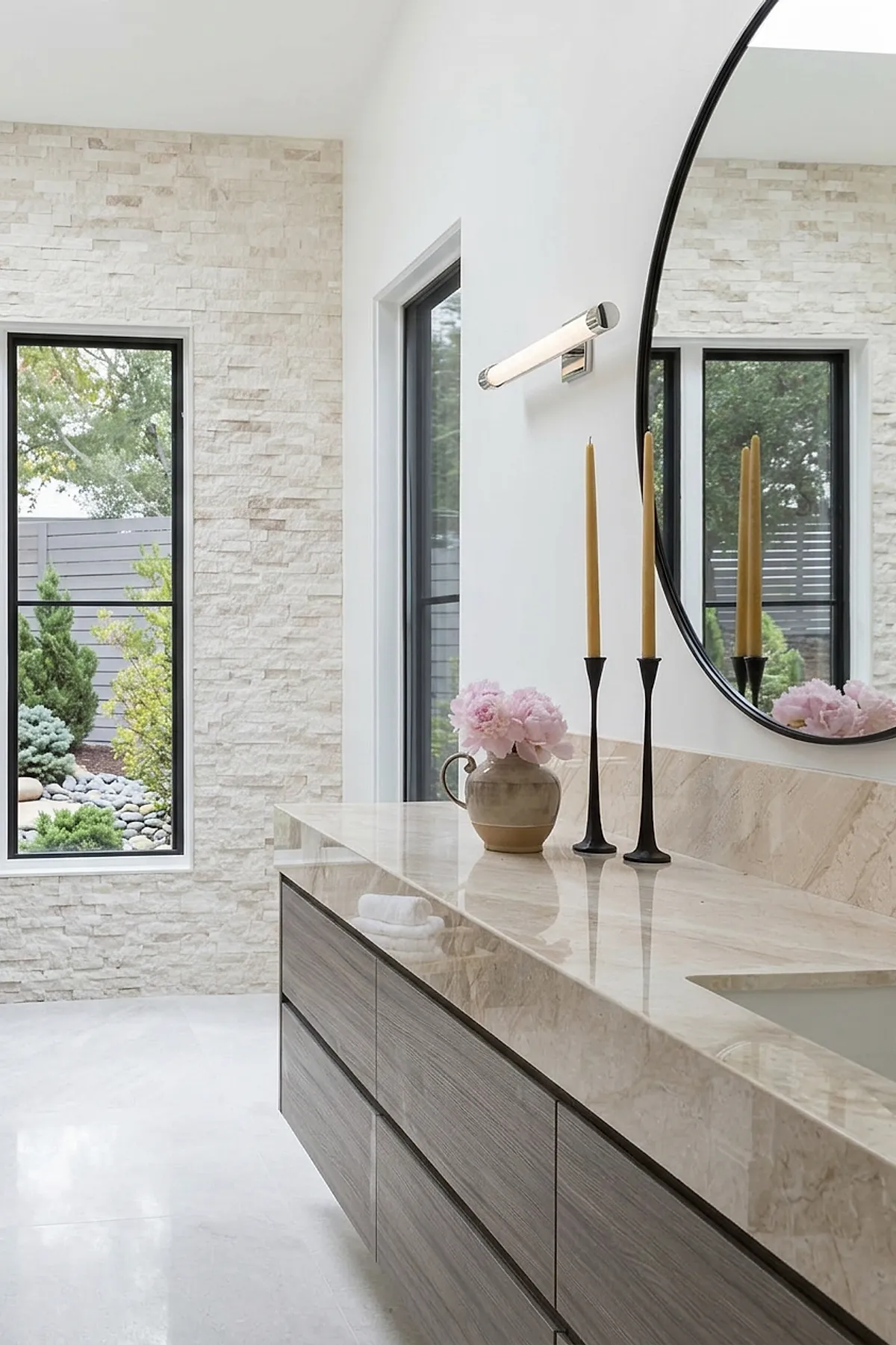 Warm neutral bathroom with a polished marble countertop vanity featuring light wood drawers, a large round black-framed mirror, two tall mustard-colored candles in black holders, and a ceramic vase with pink flowers, against textured beige stone wall and large windows showing outdoor greenery.