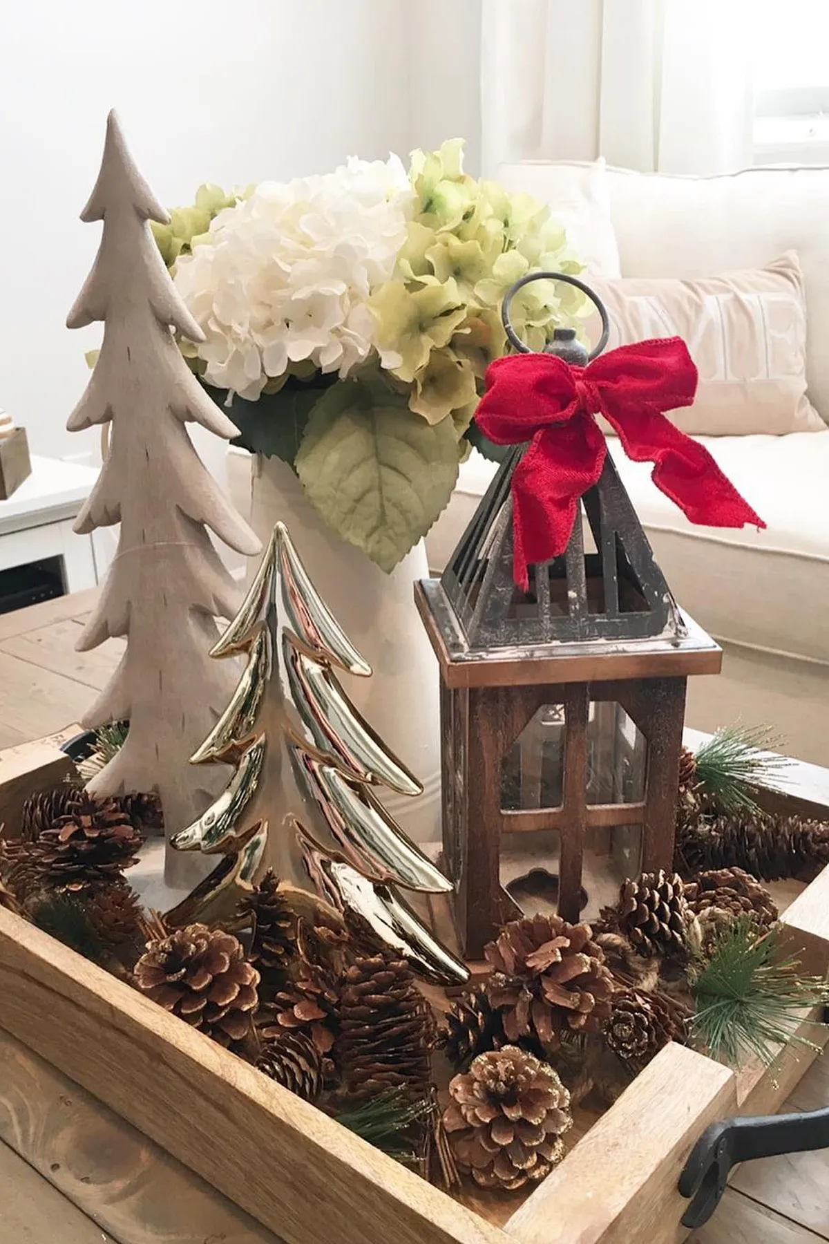 Cozy living room with wooden tray, gold tree, red bow lantern, pinecones, flowers, and a sofa featuring a LOVE cushion.