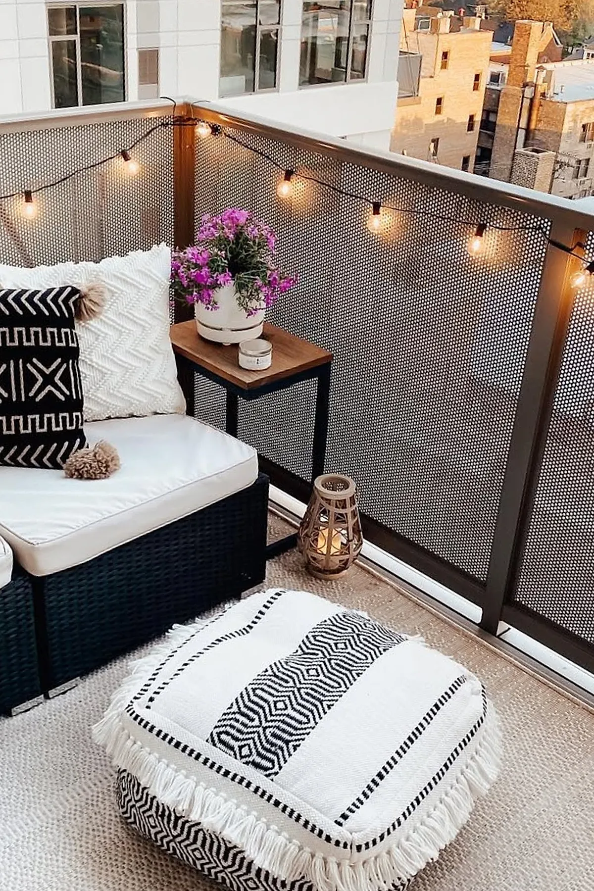 Small outdoor patio with black wicker sectional sofa featuring white cushions and textured black and white pillows, a matching black and white patterned pouf with fringe, wooden side table with a potted purple flower and candle, string lights draped along the metal railing, and a woven neutral rug on the floor.