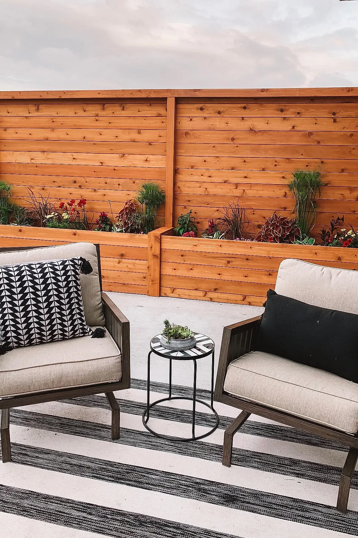 Wooden planter privacy wall with green and red plants behind cushioned patio chairs with black and white pillows, next to a small round table on a striped outdoor rug