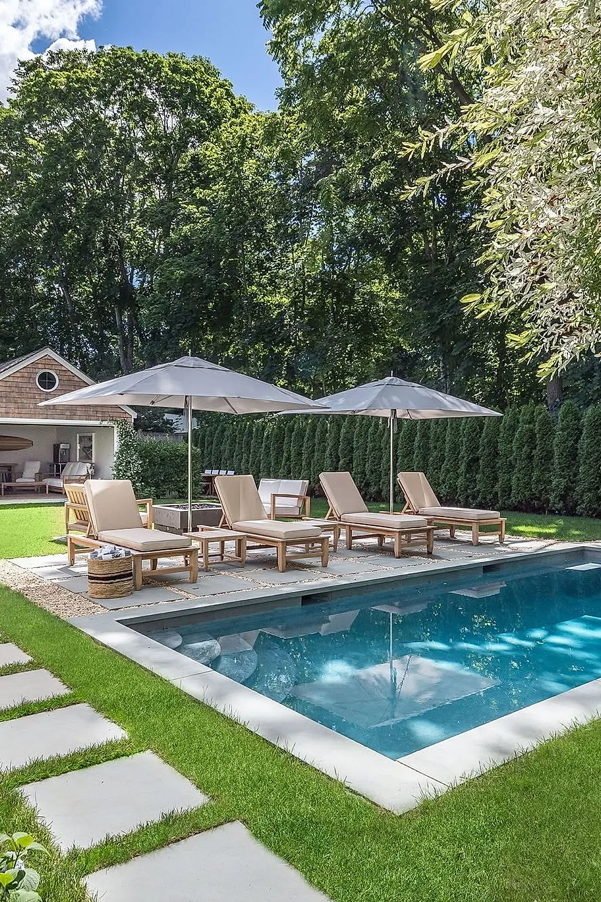 A small rectangular inground pool with clear blue water surrounded by stone pavers and green grass, accompanied by four wooden chaise lounges with beige cushions and two large white umbrellas in a backyard bordered by tall evergreen trees and a small shed.