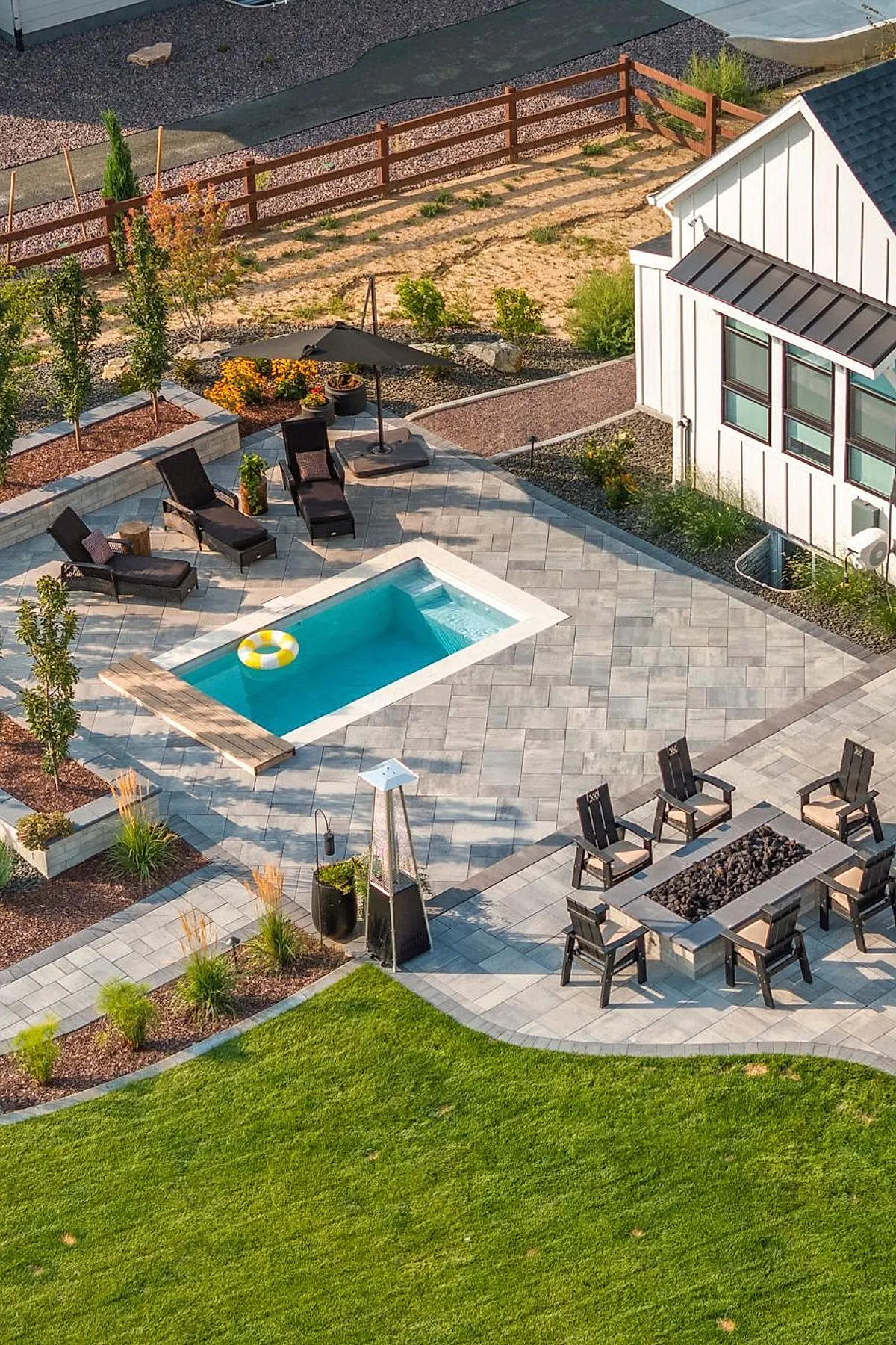 Modern rectangular plunge pool with turquoise water in a grey tiled patio surrounded by black cushioned loungers, a black umbrella, planters with trees and flowers, and a fire pit area with cushioned black chairs near a white house with black roof accents.