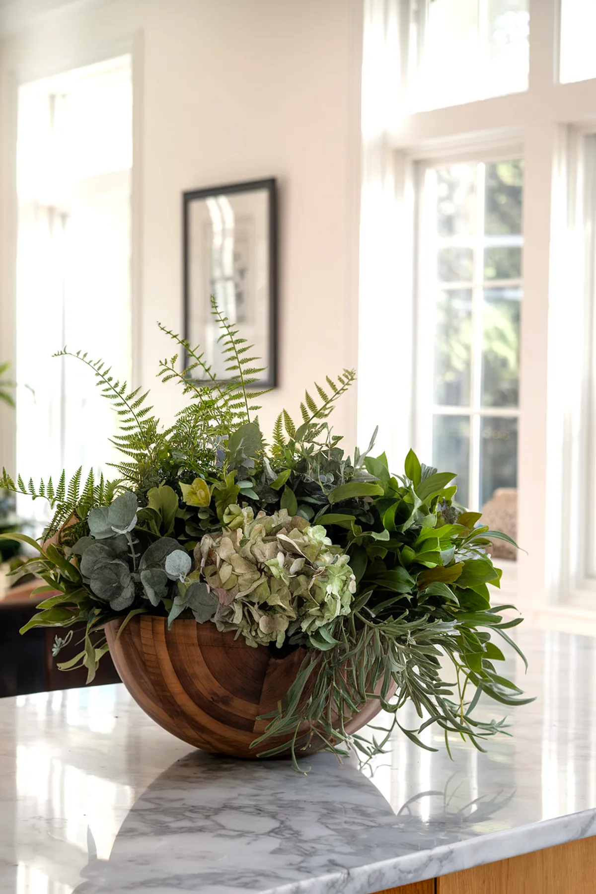 A wooden bowl filled with various green plants and flowers on a marble table.