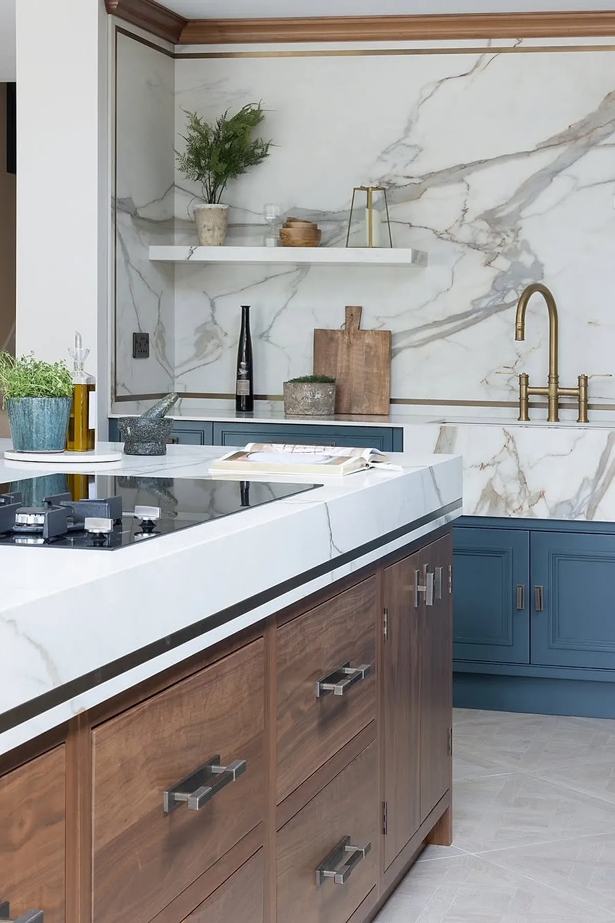 Modern kitchen featuring white marble countertops and backsplash with gray veining, blue lower cabinets with silver hardware, wooden drawer fronts on island, brass faucet, open shelf with plants and wood decor items.