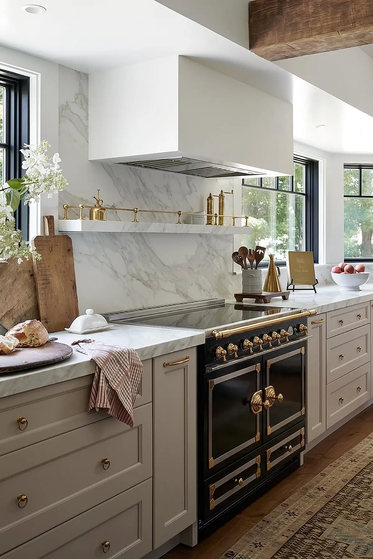 Kitchen countertop styled with white marble surface and matching backsplash, brass hardware and rail, black and gold stove, wooden cutting boards leaning against the wall, a bowl of red apples, and natural light pouring through black-framed windows.