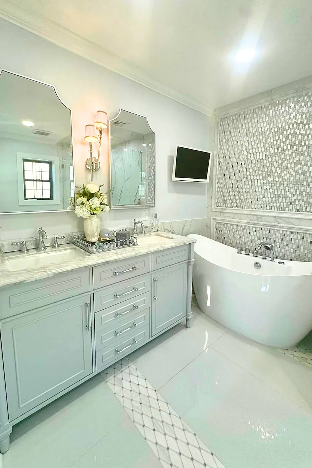 Luxury master bathroom featuring a double vanity with light gray cabinetry and marble countertop, two decorative mirrors with sconces between them, a freestanding white bathtub against a wall with mosaic tile accent, and a small wall-mounted TV.