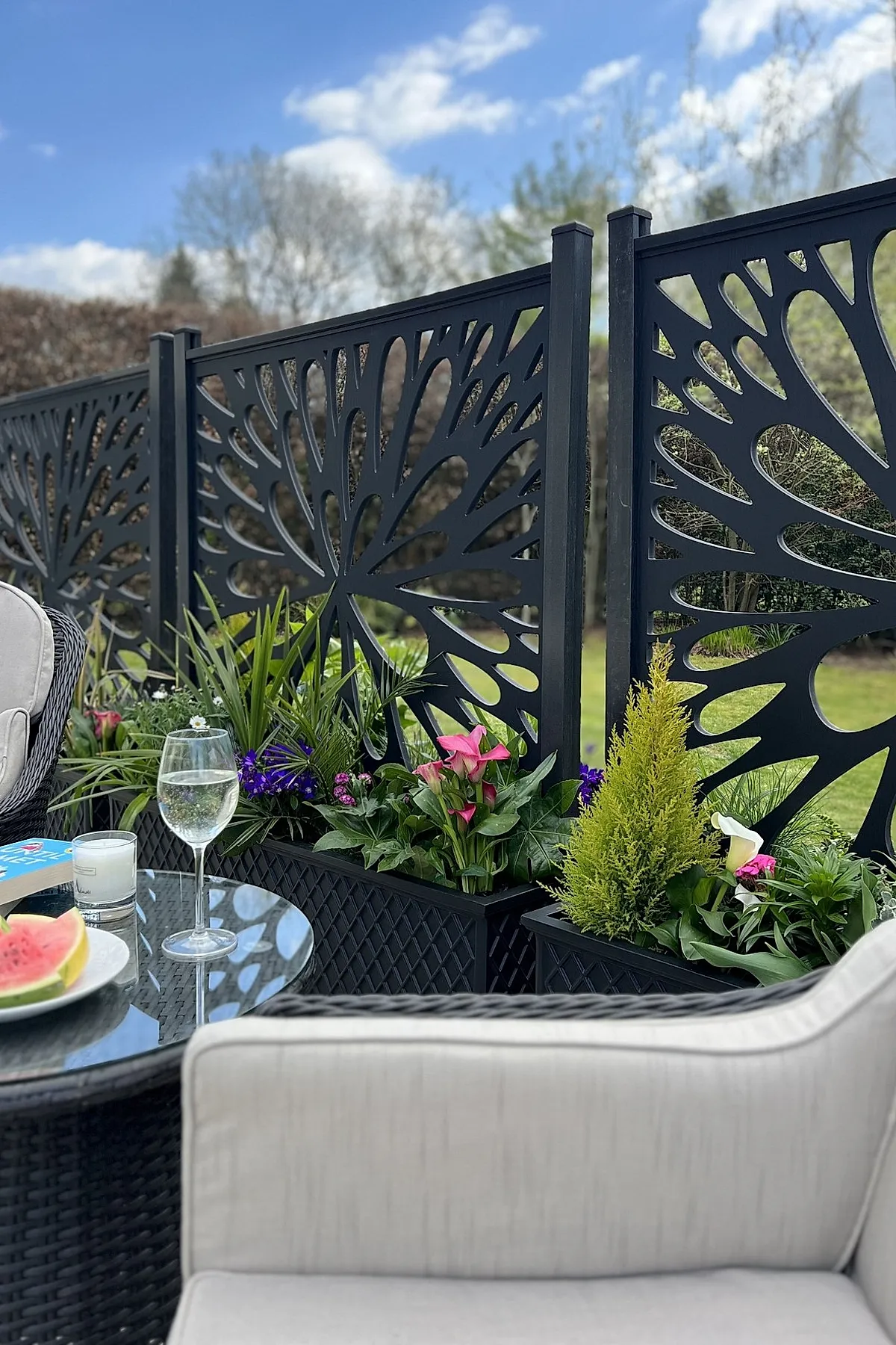 Black metal planter privacy wall with decorative floral cutouts, filled with lush green plants and colorful flowers, set beside patio seating under a blue sky