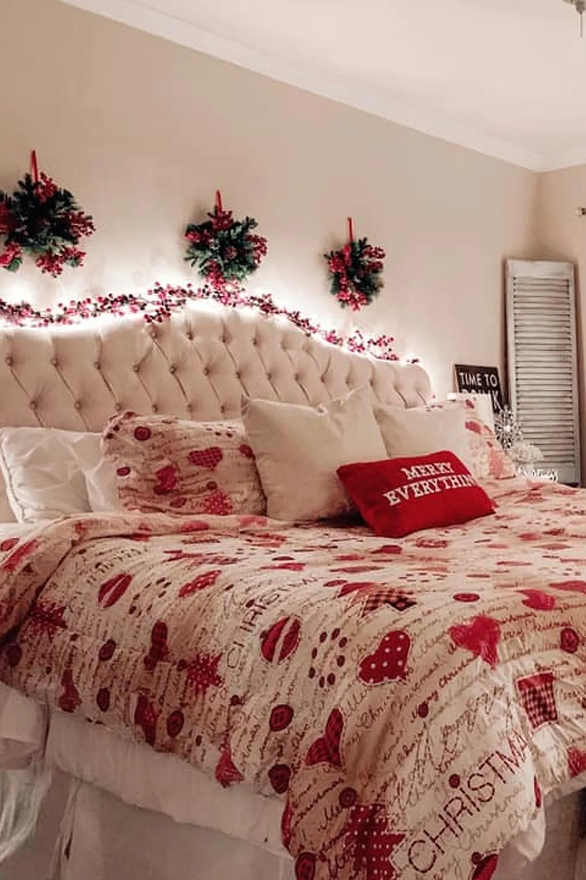 Christmas bedroom featuring a tufted beige headboard decorated with string lights and red berry garlands, three green wreaths with red ribbons hanging above, bedding with red and beige holiday-themed patterns including hearts and stars, assorted pillows including a red one that says
