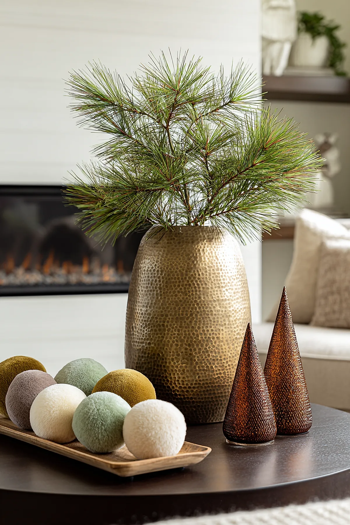 christmas coffee table decor featuring a large hammered bronze vase filled with long green pine branches, accompanied by a wooden tray holding eight soft textured balls in shades of cream, sage green, mustard yellow, and taupe, alongside two brown textured cone-shaped decorative trees on a dark round coffee table with fireplace and neutral furniture in the background.