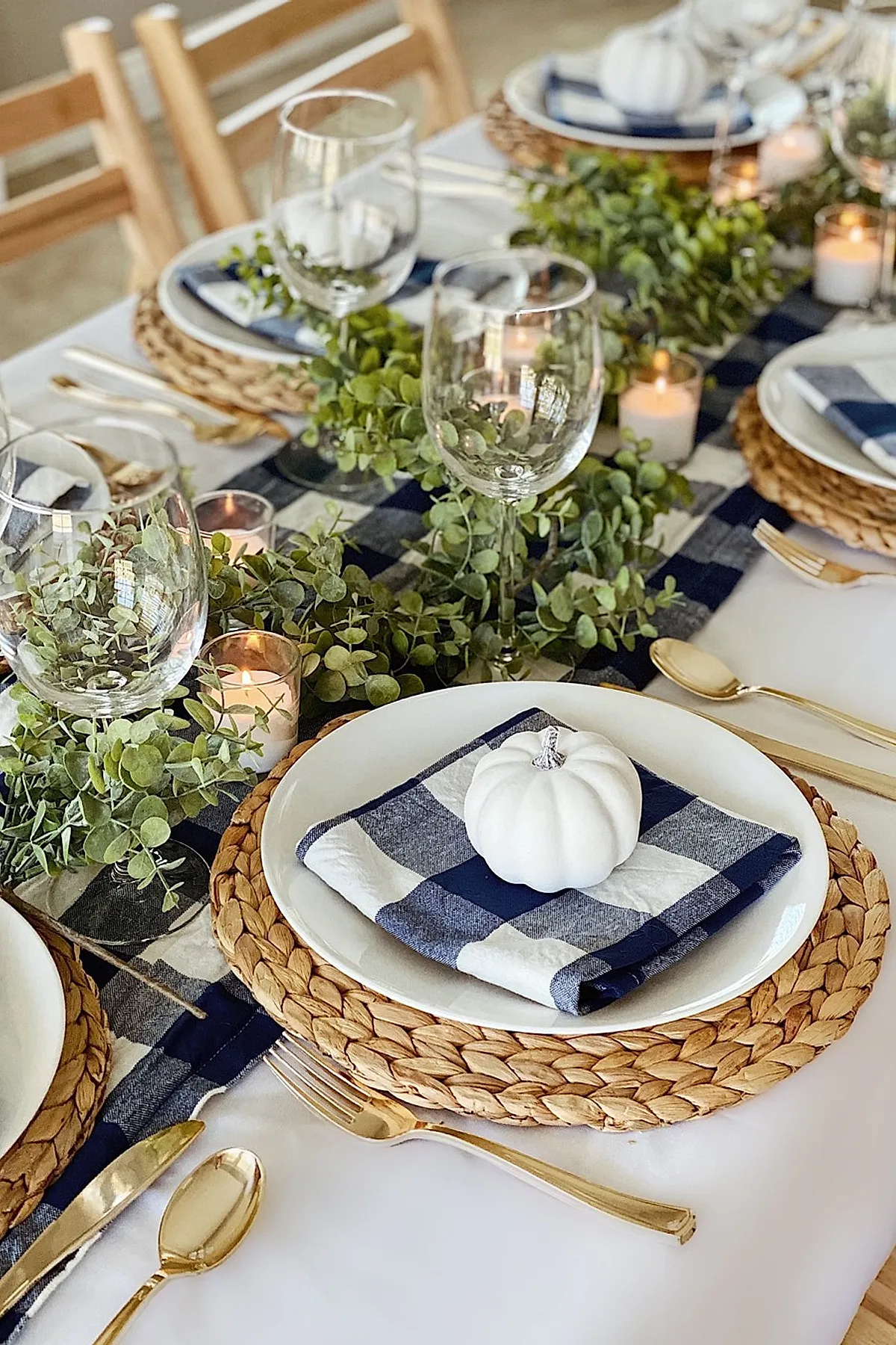 Thanksgiving table setting with blue and white checkered fabric runner, woven chargers, white plates topped with blue and white checkered napkins and small white pumpkins, surrounded by eucalyptus garland and lit votive candles