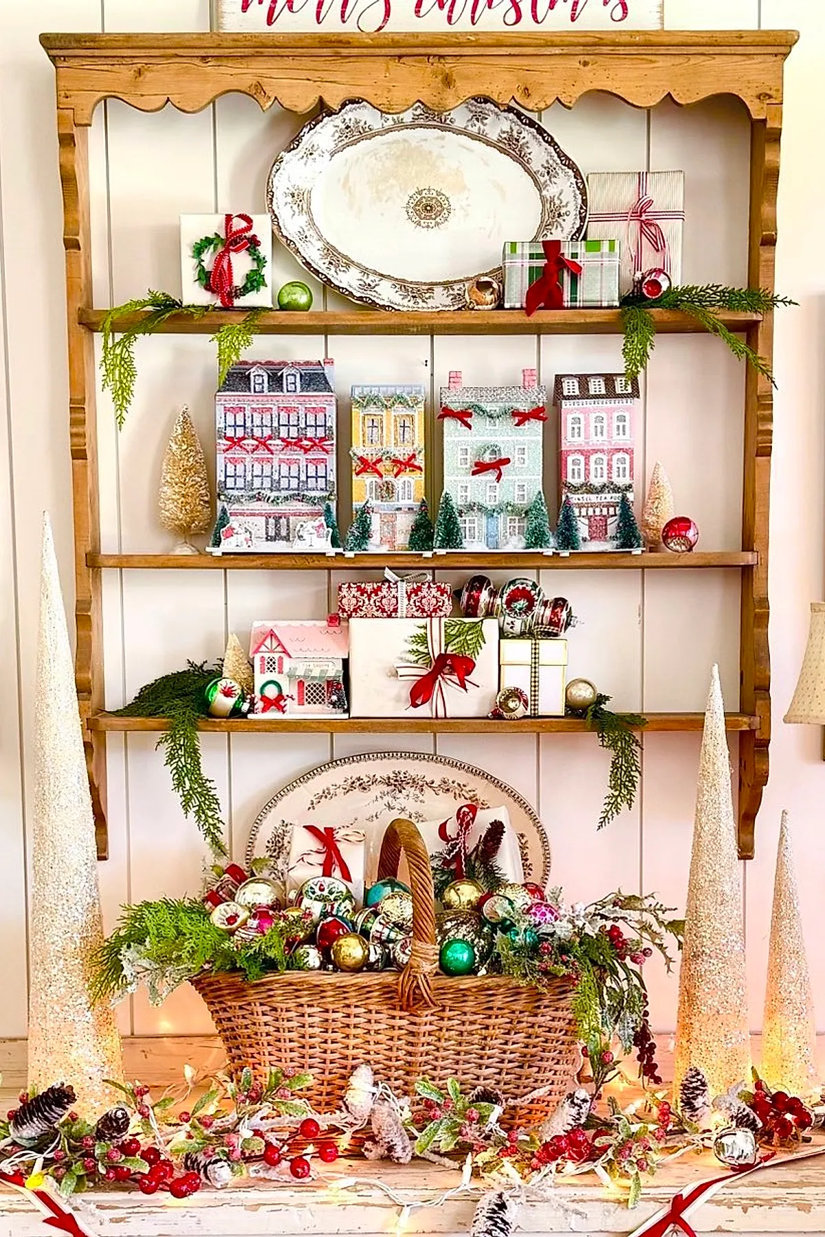 Wooden shelves decorated with a miniature village of pastel houses, bottle brush trees, and colorful glass ornaments for a nostalgic vintage christmas aesthetic.