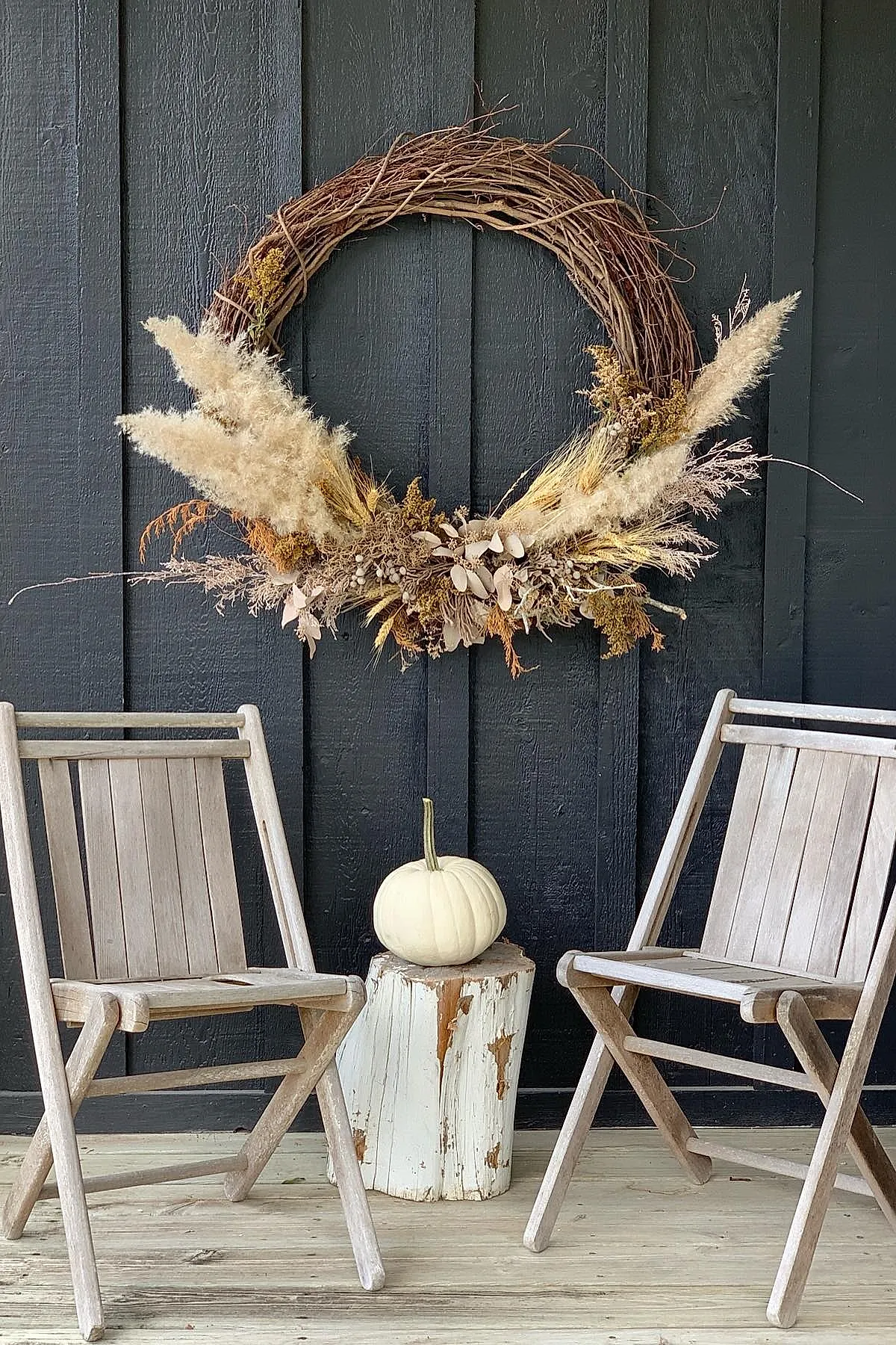 wreath made of dried pampas grass, wheat, and wildflowers arranged asymmetrically on a brown grapevine base hanging on a black painted vertical wood panel wall above two rustic wooden chairs with weathered finish and a white pumpkin placed on a worn white tree stump between them
