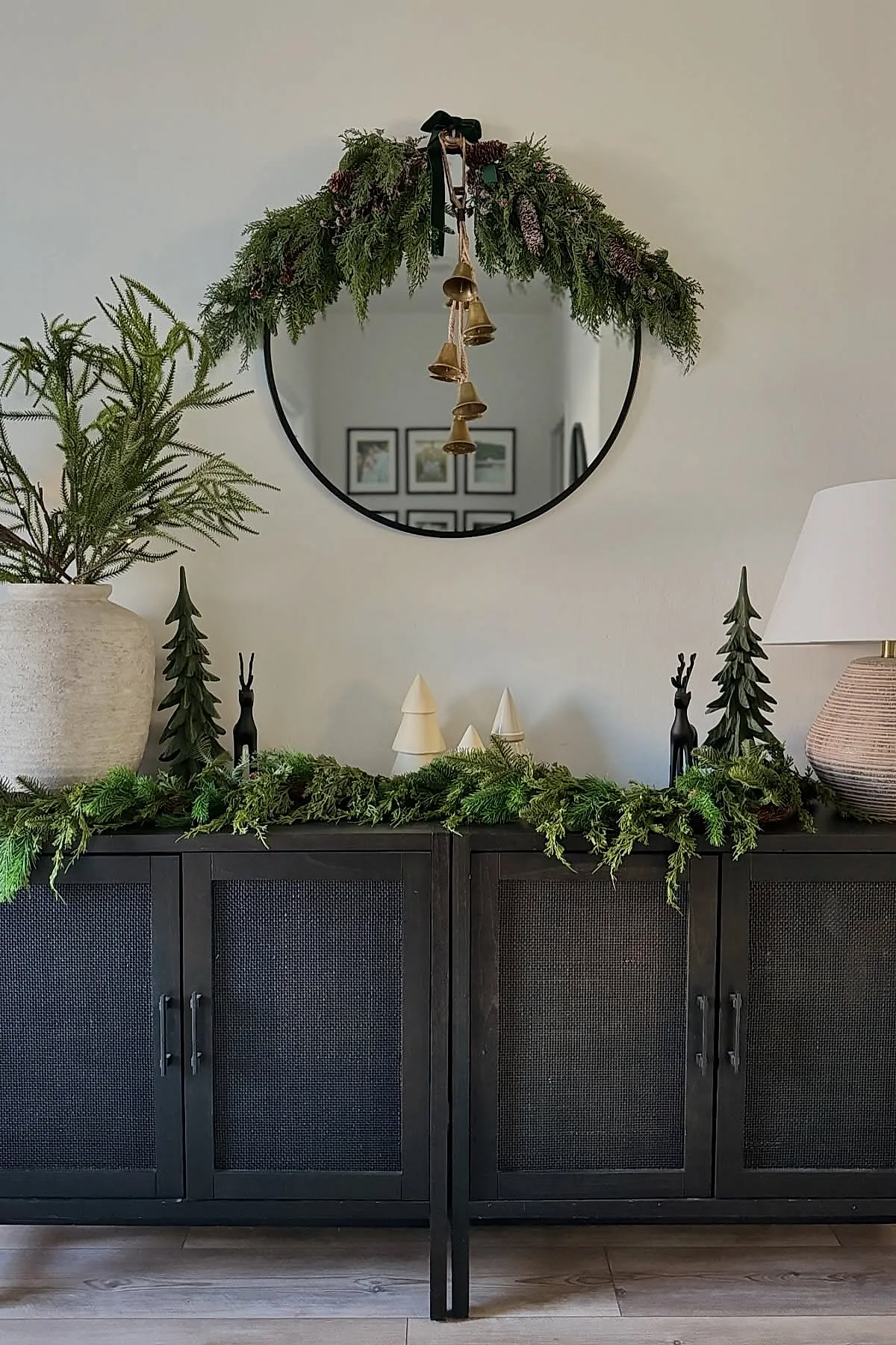 minimalist christmas decor with green garland and pine branches draped on a black cabinet and round mirror, accented by hanging gold bells, white ceramic trees, black reindeer figurines, and a large textured vase with greenery