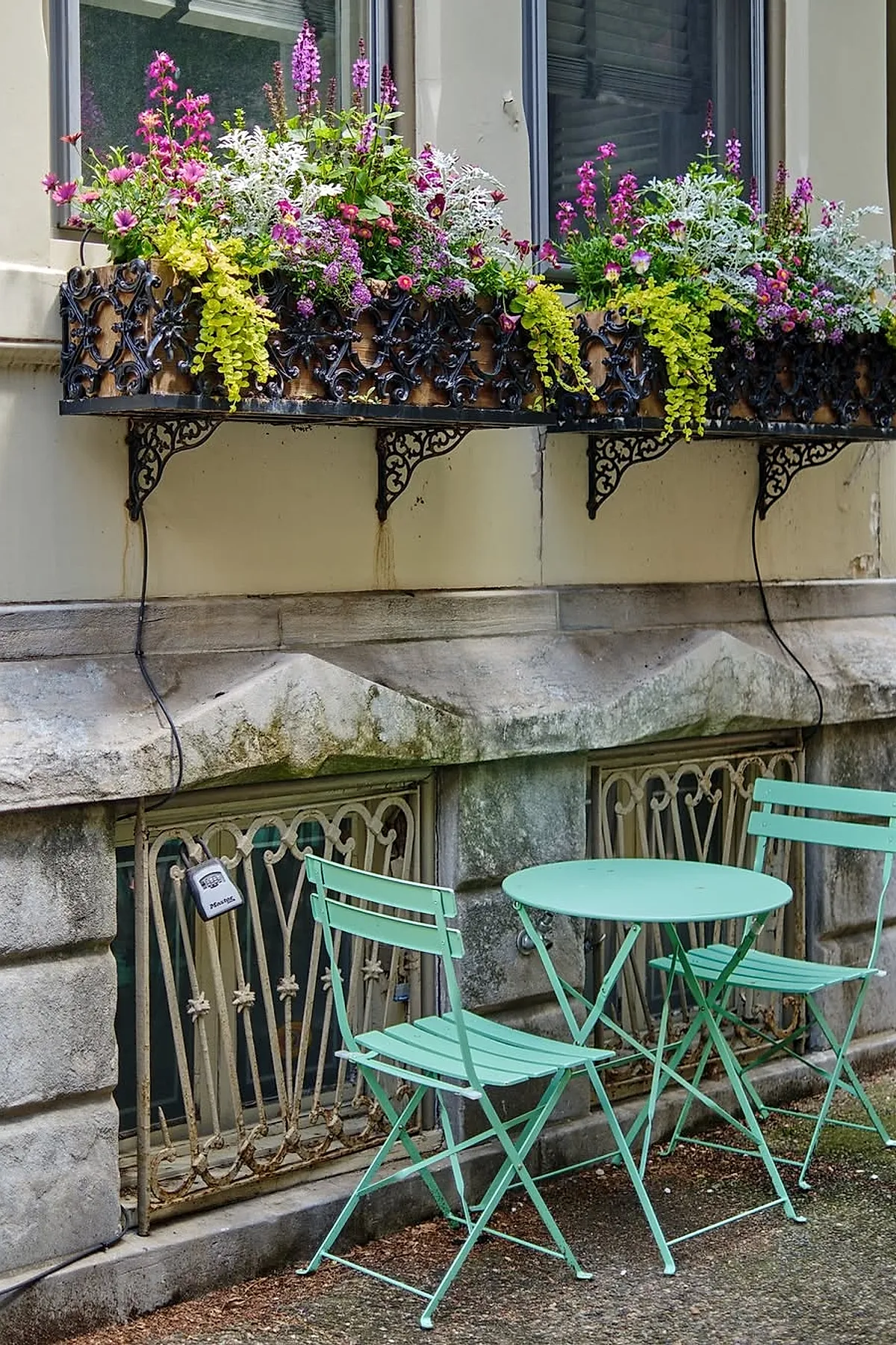 Ornate black iron window box overflowing with pink, white, and purple flowers and yellow trailing foliage mounted under two windows on a cream building wall with a matching mint green metal bistro table and two chairs on the stone pavement below.
