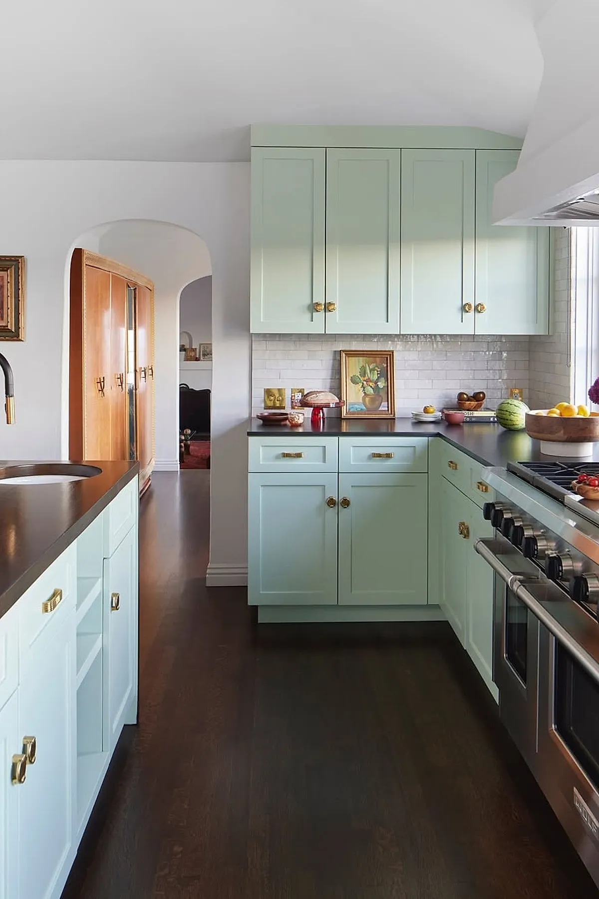 Modern Spanish style kitchen featuring soft green cabinetry with brass hardware, dark wood floors, white subway tile backsplash, black countertops, stainless steel stove, and an arched doorway leading to another room.