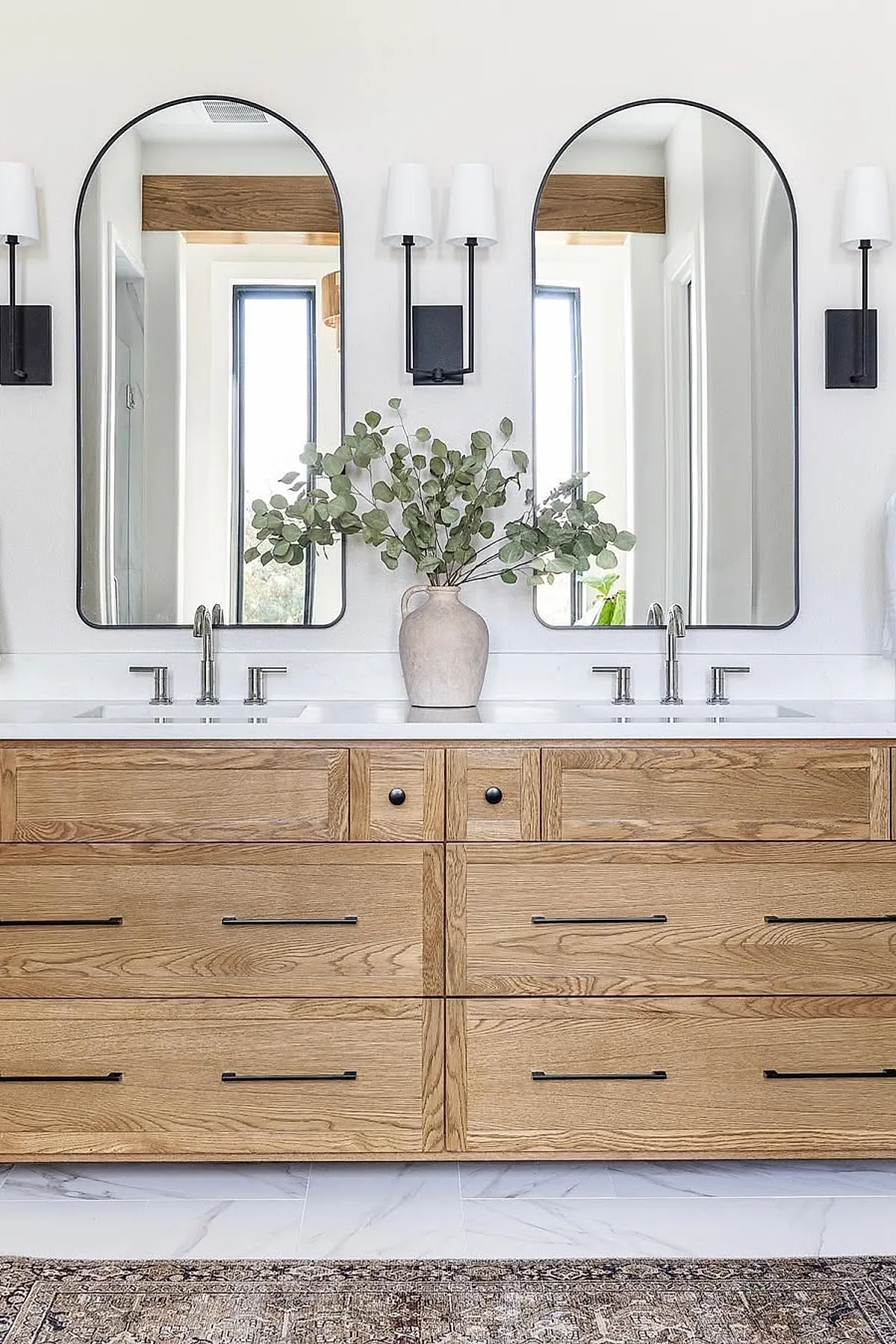 Modern master bathroom featuring a large wooden vanity with black handles, two tall arched black-framed mirrors above dual sinks, white walls, silver faucets, black wall sconces with white shades, and a beige vase holding green eucalyptus branches on the countertop.
