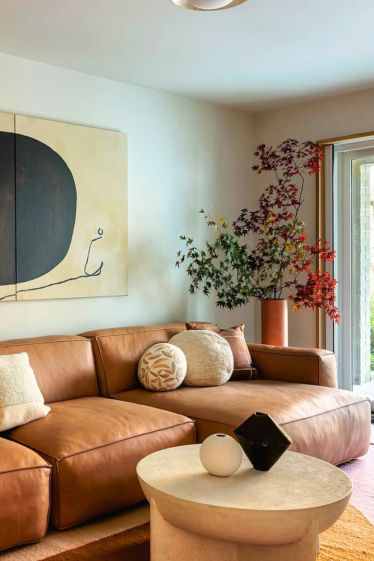 Living room featuring a cognac brown leather sectional couch with round and textured pillows, an abstract black and beige painting on the wall, a light stone coffee table with minimalist vases, and a tall vase with green and red leafy branches near sliding glass doors.