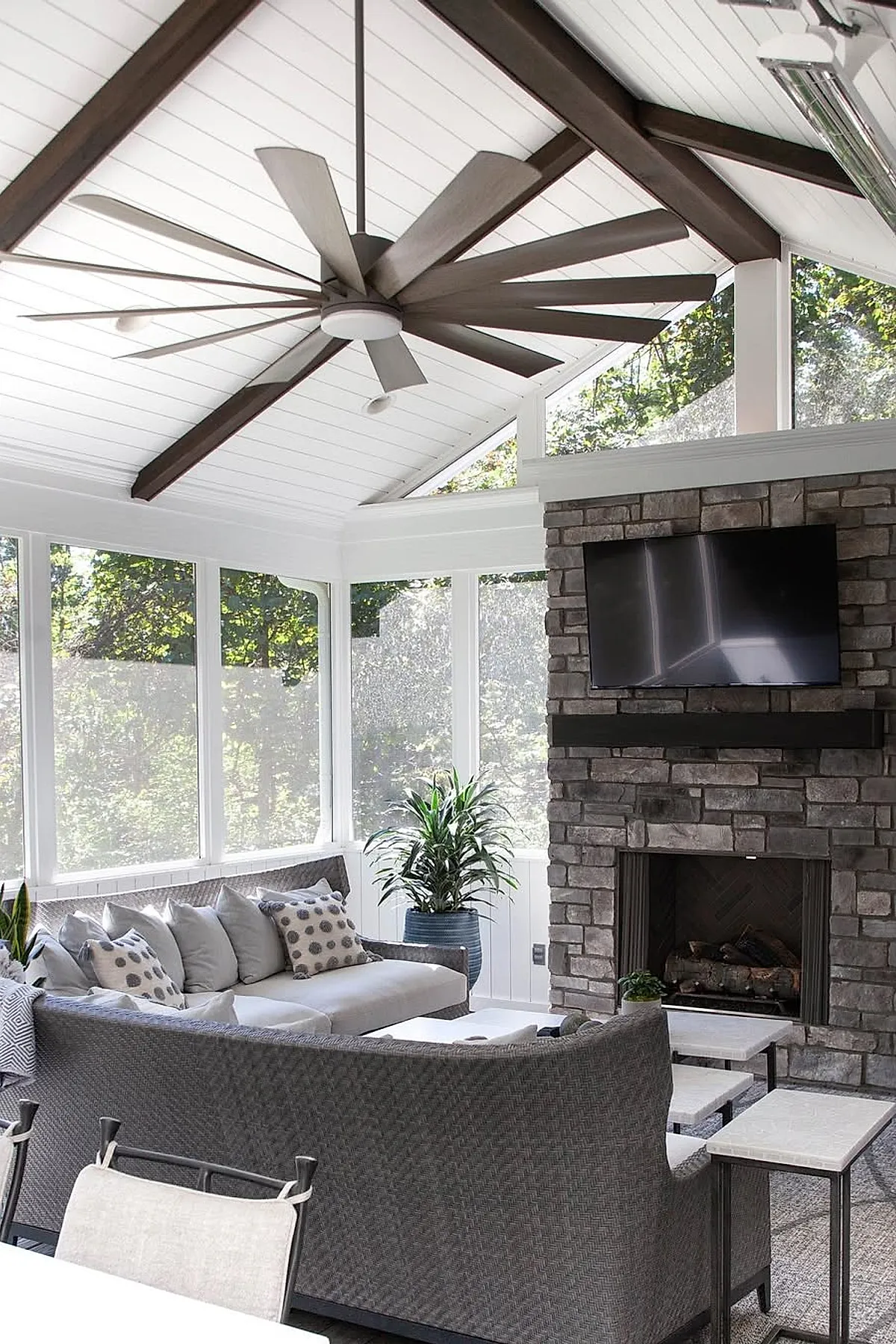Screened in porch featuring stone fireplace with mounted TV above, large woven sectional sofa with neutral cushions and patterned pillows, white ceiling with dark wood beams and large ceiling fan, abundant natural light from screened windows overlooking greenery