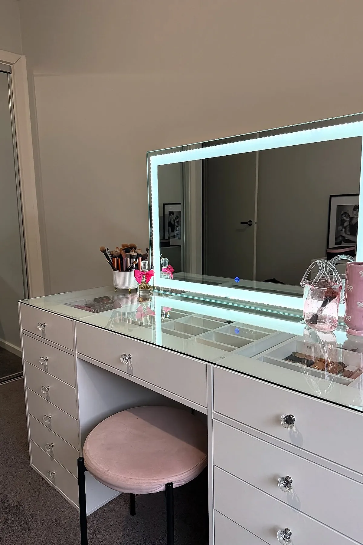 white bedroom vanity with glass top revealing organized storage underneath, large rectangular LED-lit mirror, round pink cushioned stool with black legs, makeup brushes in white container, pink decorative jar on desk