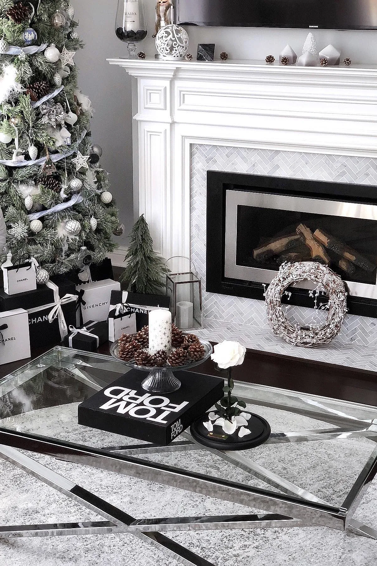 modern christmas coffee table decor featuring a large white candle surrounded by pinecones on a glass cake stand, placed on top of a black Tom Ford book on a reflective metal and glass coffee table, next to a single white rose in a small vase, with a decorated Christmas tree and fireplace in the background.