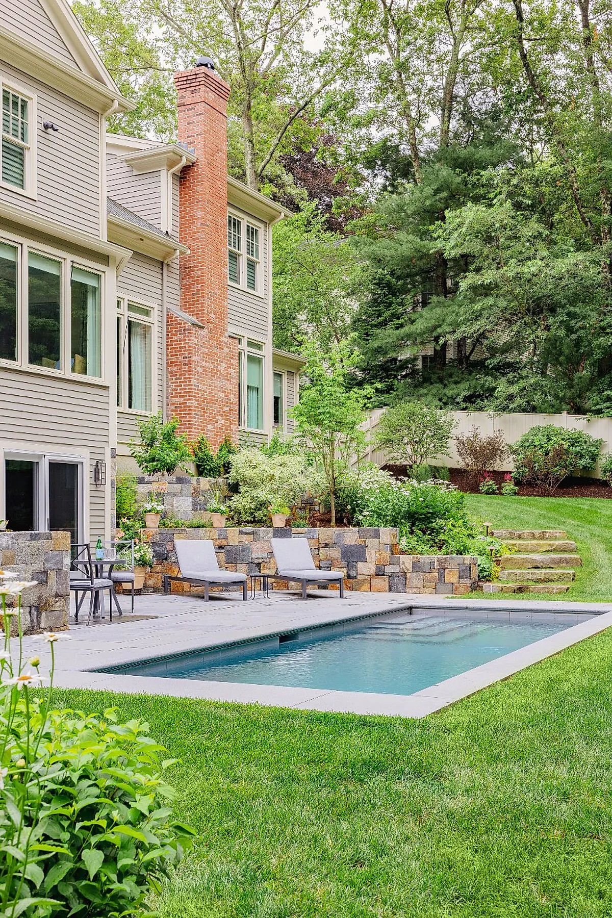 Rectangular plunge pool with clear water bordered by smooth gray concrete, flanked by multi-colored stacked stone retaining walls with potted plants, adjacent to green grass lawn and lush garden shrubs in front of a beige house with large windows and red brick chimney.