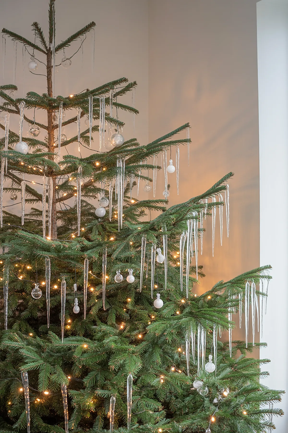 Natural green Christmas tree decorated with long clear icicle ornaments, white round baubles, and warm white string lights against a neutral wall background.