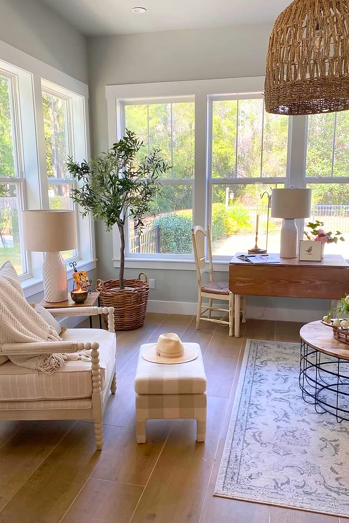 Sunroom with large windows showing greenery outside, light beige armchair with a textured throw, matching ottoman topped with a straw hat, wicker basket planter with tall green plant, wooden desk with lamp and chair, round wooden coffee table on patterned rug, and a large woven pendant light overhead.