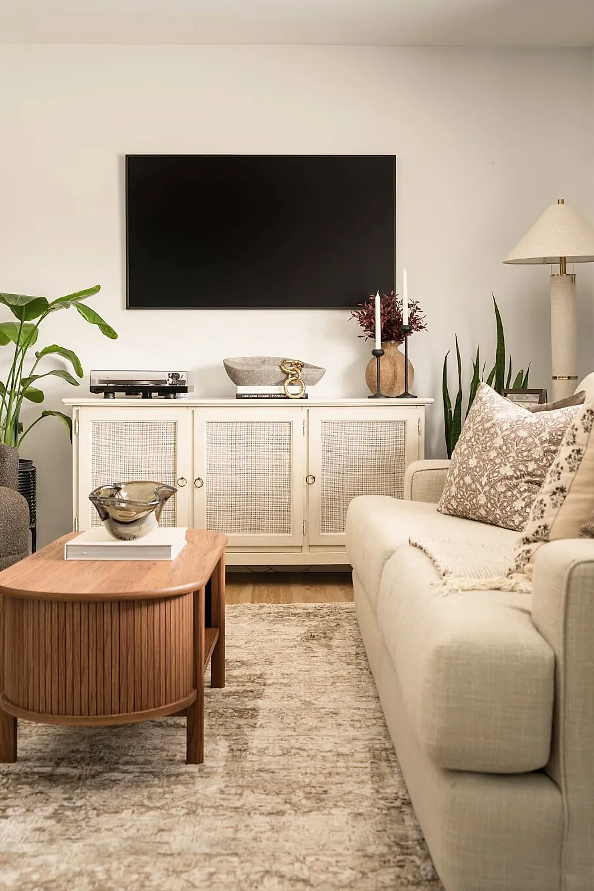 Earthy living room featuring a light beige sofa with patterned throw pillows and a cream throw blanket, a wooden coffee table with curved edges topped with books and a decorative bowl, a white media console with rattan doors holding decorative items, green leafy plants on both sides, and a floor lamp with a beige shade against off-white walls.