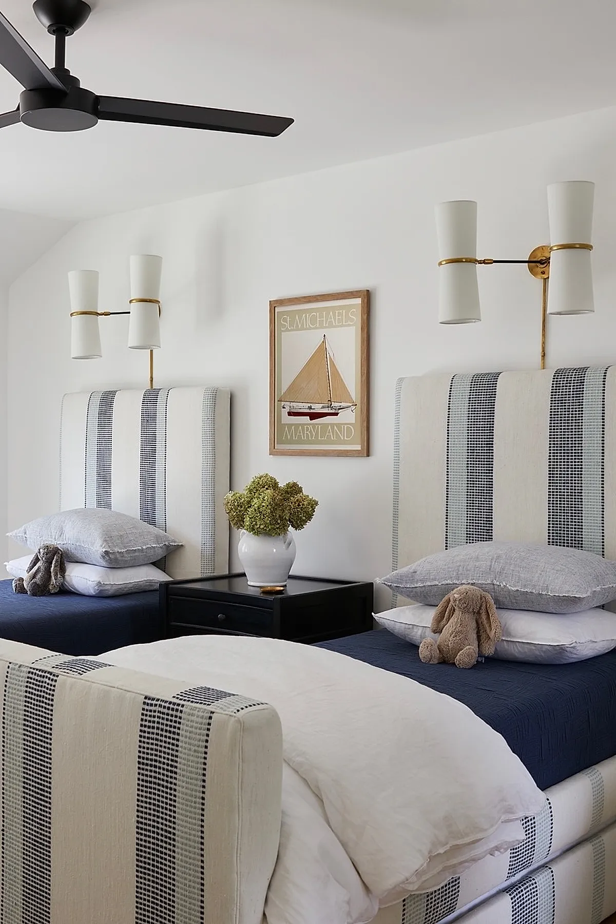 Two twin beds with striped upholstered headboards in cream and black, navy blue bedding, stacked gray and white pillows, brown plush bunny toys on each bed, black nightstand with white vase holding dried flowers, white walls with framed sailboat print.