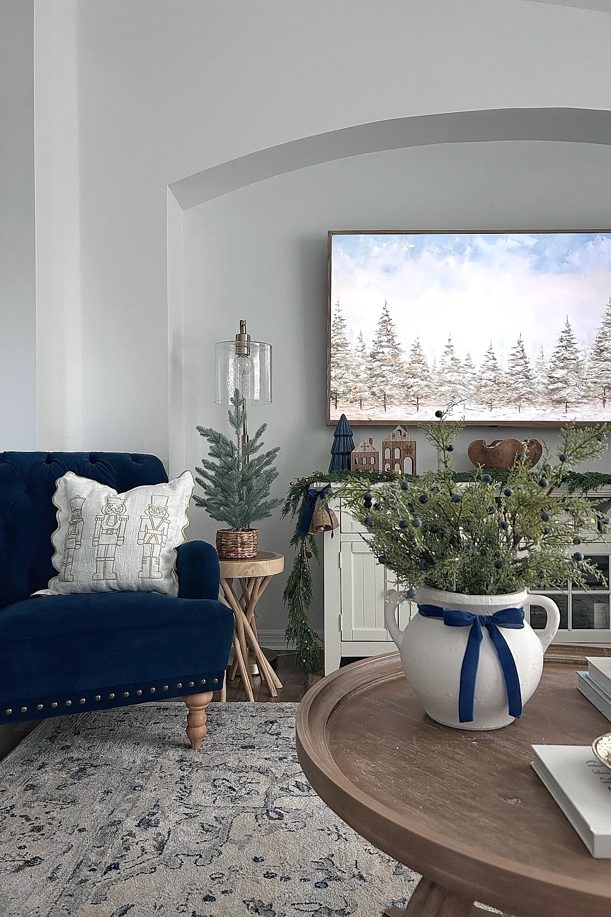 christmas coffee table decor featuring a white ceramic pitcher tied with a navy blue ribbon holding greenery with dark berries, placed on a round wooden coffee table with books, alongside a navy blue velvet armchair with nutcracker pillow, small tabletop pine tree on wooden side table, and winter-themed artwork in the background.