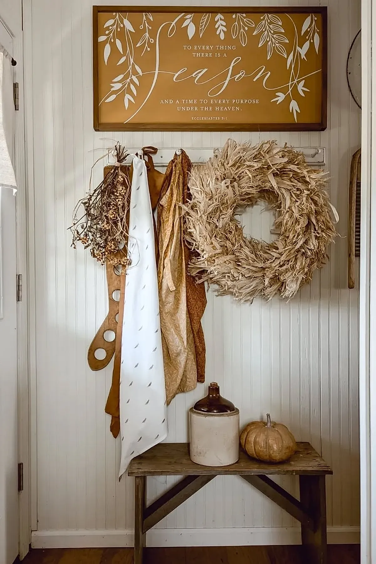 farmhouse entryway with white beadboard walls, neutral tan wreath, dried branches, hanging scarves in cream and mustard tones, rustic bench with ceramic jug and small pumpkin