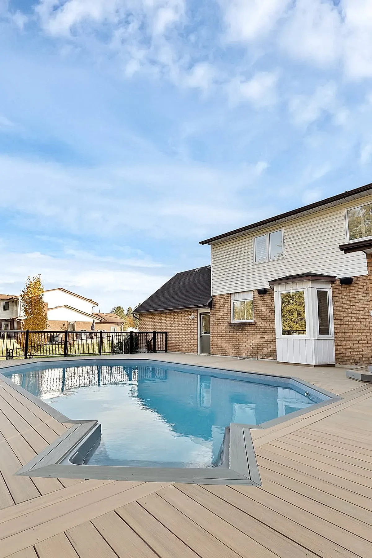 A backyard pool with a light beige composite deck surrounding a rectangular swimming pool next to a two-story brick and siding house under a partly cloudy sky.