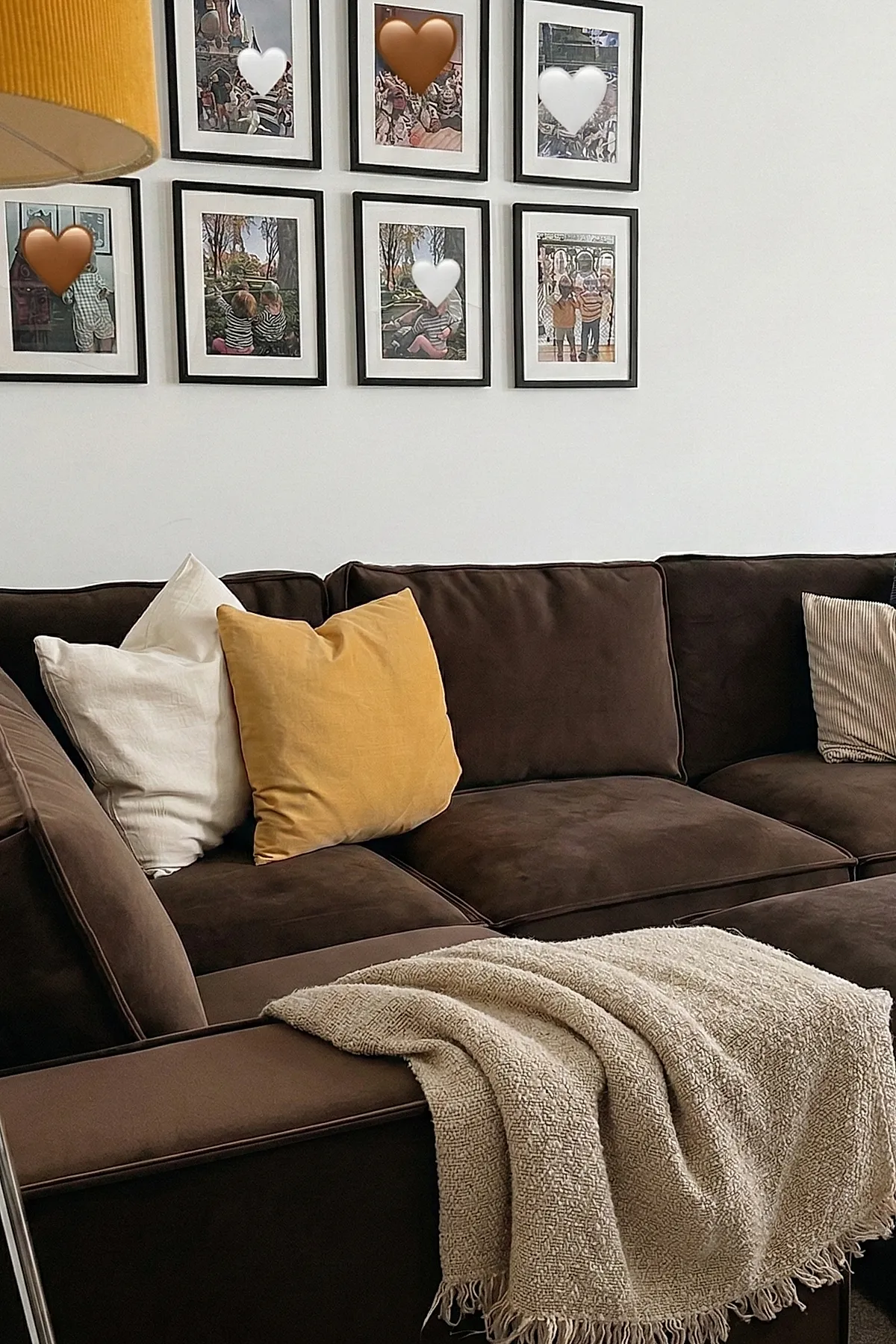 Chocolate brown sofa adorned with cream, mustard yellow, and beige striped pillows, draped with a textured cream throw blanket. The background features a white wall decorated with a gallery of framed photos.
