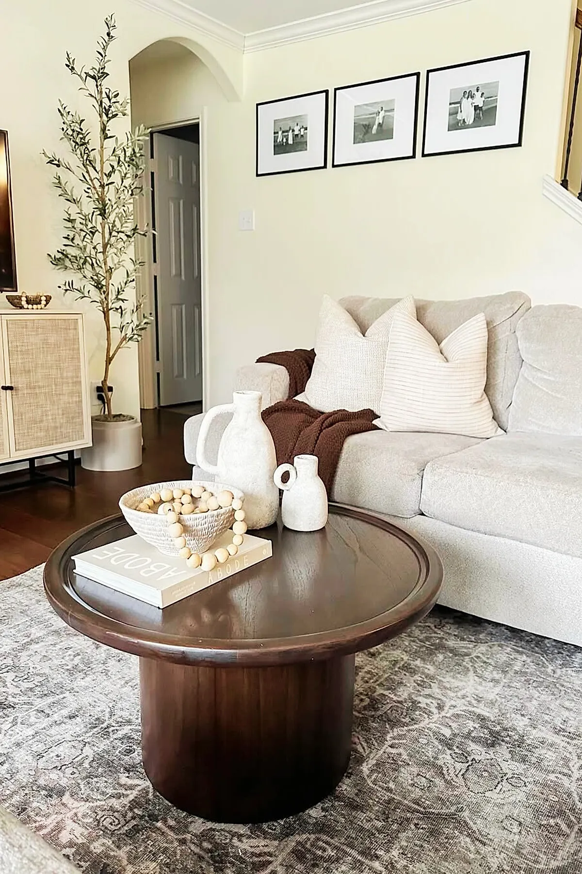 Warm earthy living room featuring a dark wooden round coffee table topped with white ceramic pitchers, a bowl with wooden beads, and a book. A light beige sofa with neutral textured pillows and a brown throw blanket sits behind the table. The room includes a tall potted plant, light walls, framed black-and-white photos on the wall, and a patterned gray rug.