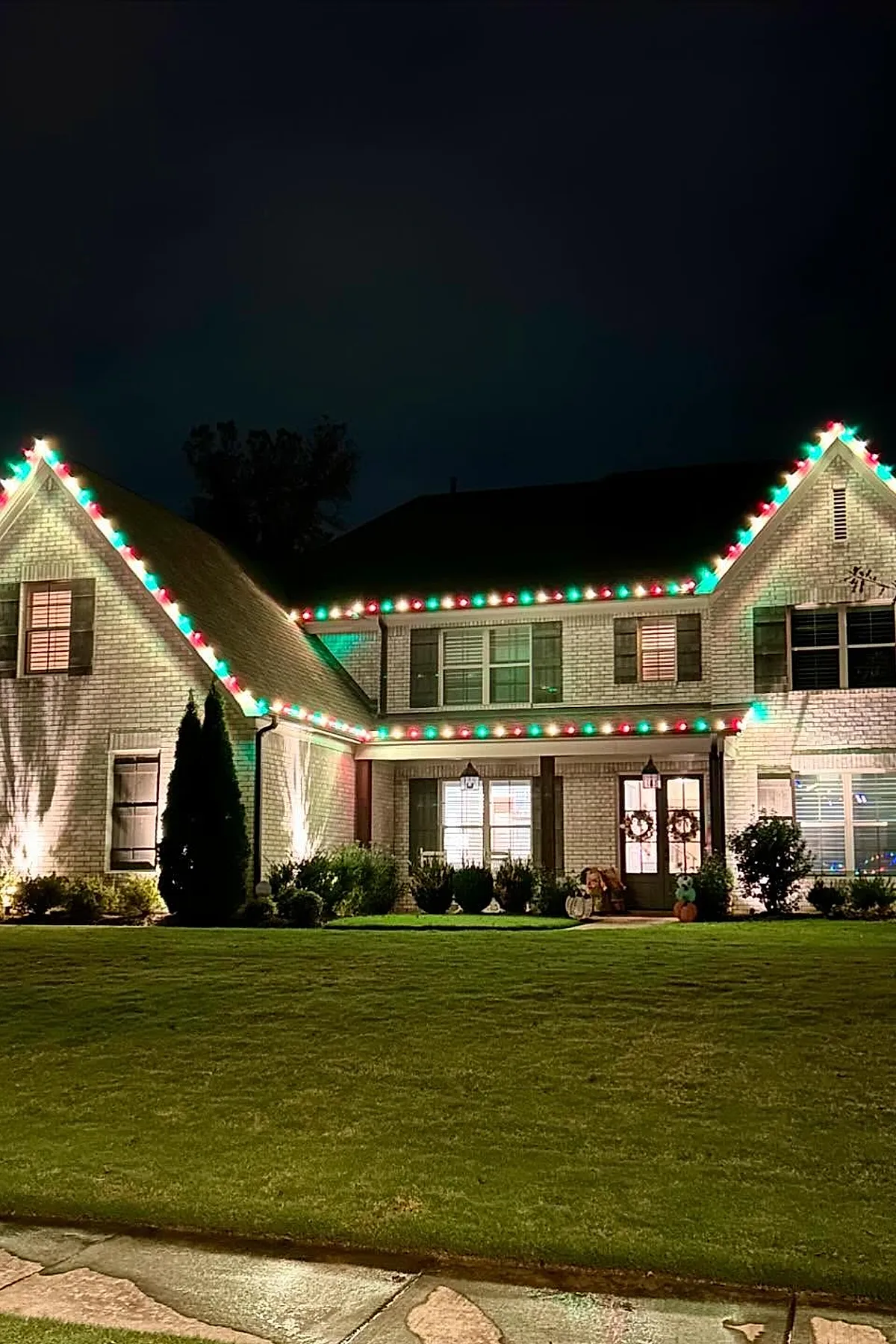 Two-story light-colored brick house decorated with red, green, and white christmas lights outlining the roof peaks and front porch at night, with lit wreaths on doors and windows and a well-kept green lawn in front.