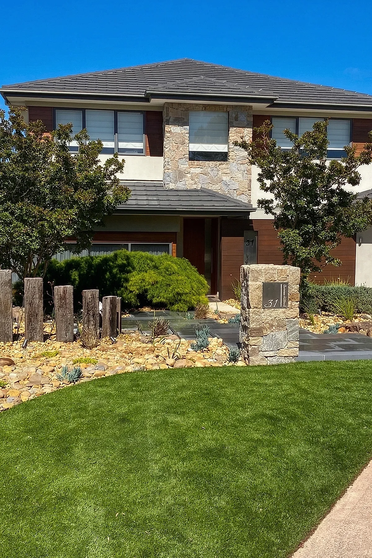 Small front yard landscaping featuring a bed of tan river rocks with scattered succulents, tall weathered wooden posts arranged vertically, lush green shrubs, two leafy trees on either side, and a stone pillar displaying the house number in front of a modern two-story home with stone and wood siding.