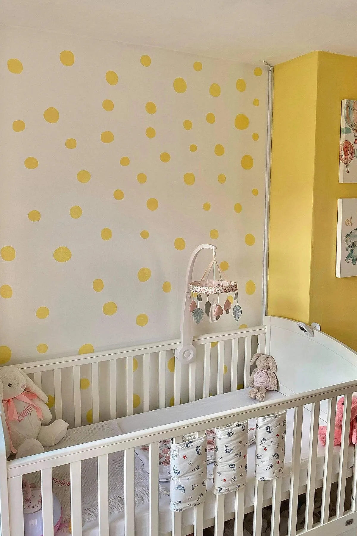 Nursery with white crib against white wall featuring scattered yellow polka dots, soft plush toys inside crib, pale yellow accent wall corner.