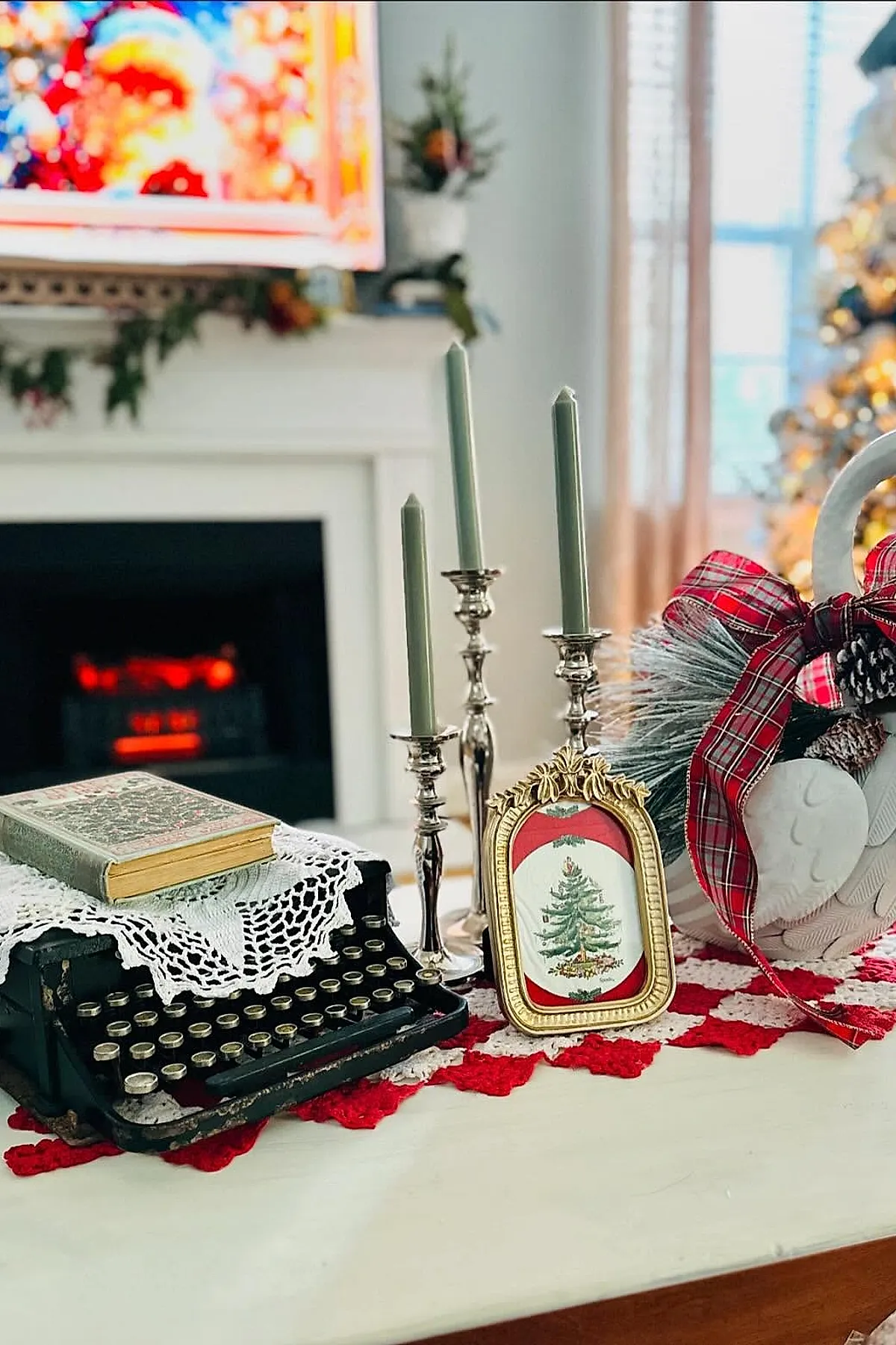 christmas coffee table decor featuring an antique black typewriter covered partially by a white crocheted cloth, a vintage book on top, three silver candlesticks with sage green taper candles, a small ornate gold frame with a Christmas tree illustration, and a swan-shaped ceramic pitcher decorated with red plaid ribbon and pinecones, arranged on a white surface with red crocheted runner.