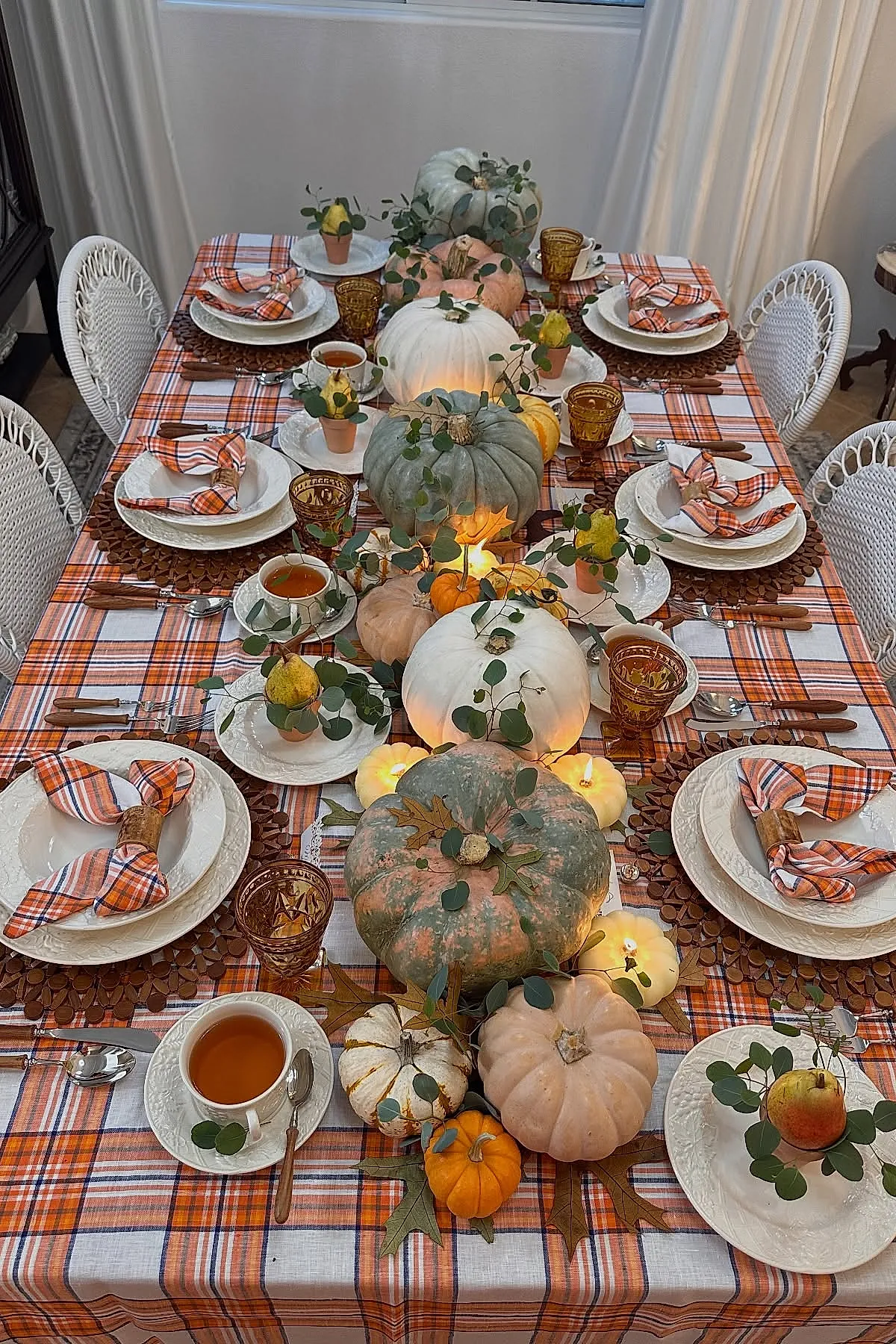 Thanksgiving table with orange plaid tablecloth, white textured plates, plaid napkins in wooden rings, amber glasses, centerpiece of white, green and pink pumpkins with eucalyptus and small pears on plates