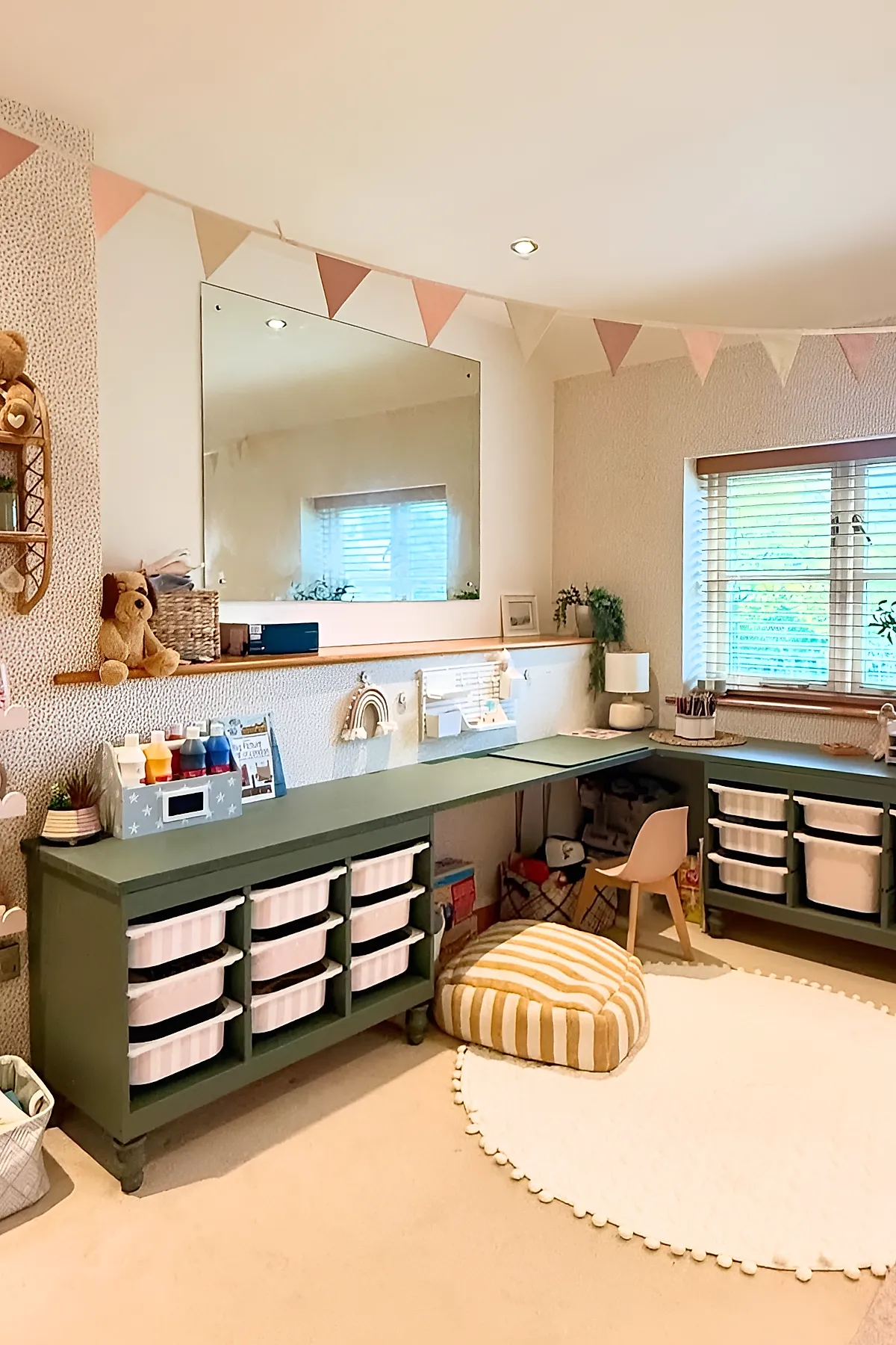 Kids desk area in bedroom with olive green built-in desks featuring multiple white storage bins, a small pink chair, beige carpet with round white rug with pom-pom trim, pastel pennant banner above, and a window with wooden blinds letting in natural light.